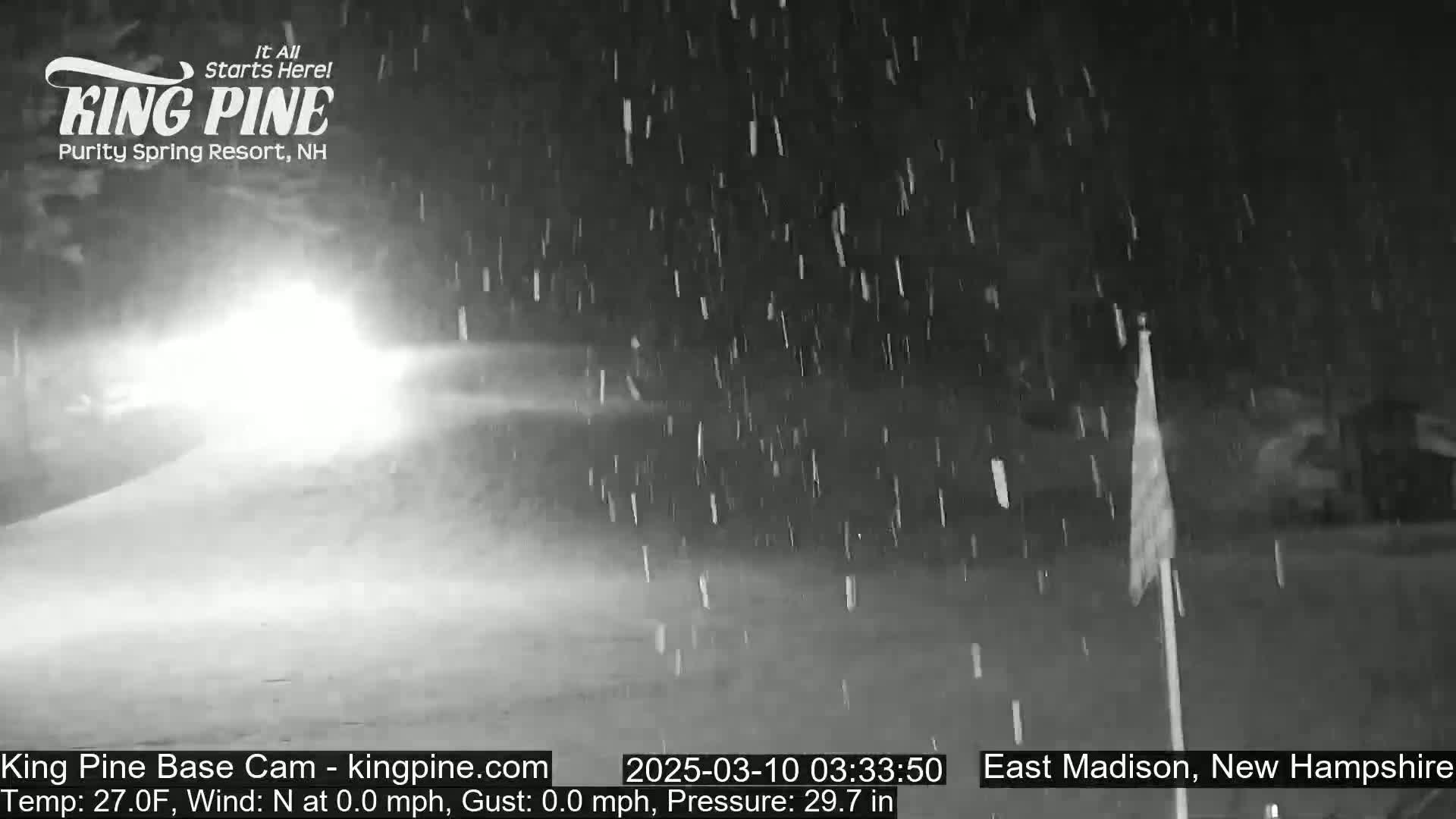 A nighttime scene of snow falling, illuminated by vehicle headlights in the distance.