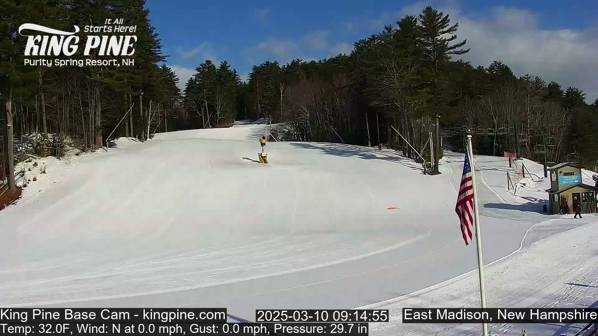 A snow-covered ski slope is visible on a sunny day, with a few skiers present and a small building at the bottom of the hill.