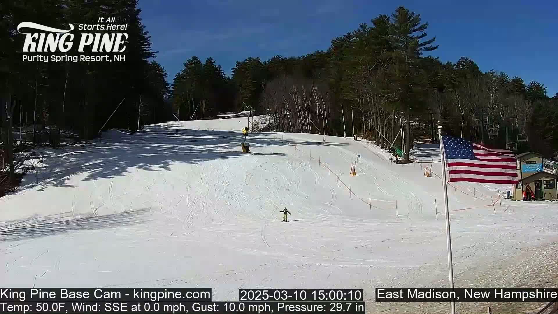 A snow-covered ski slope with a few skiers, surrounded by trees under a clear blue sky.