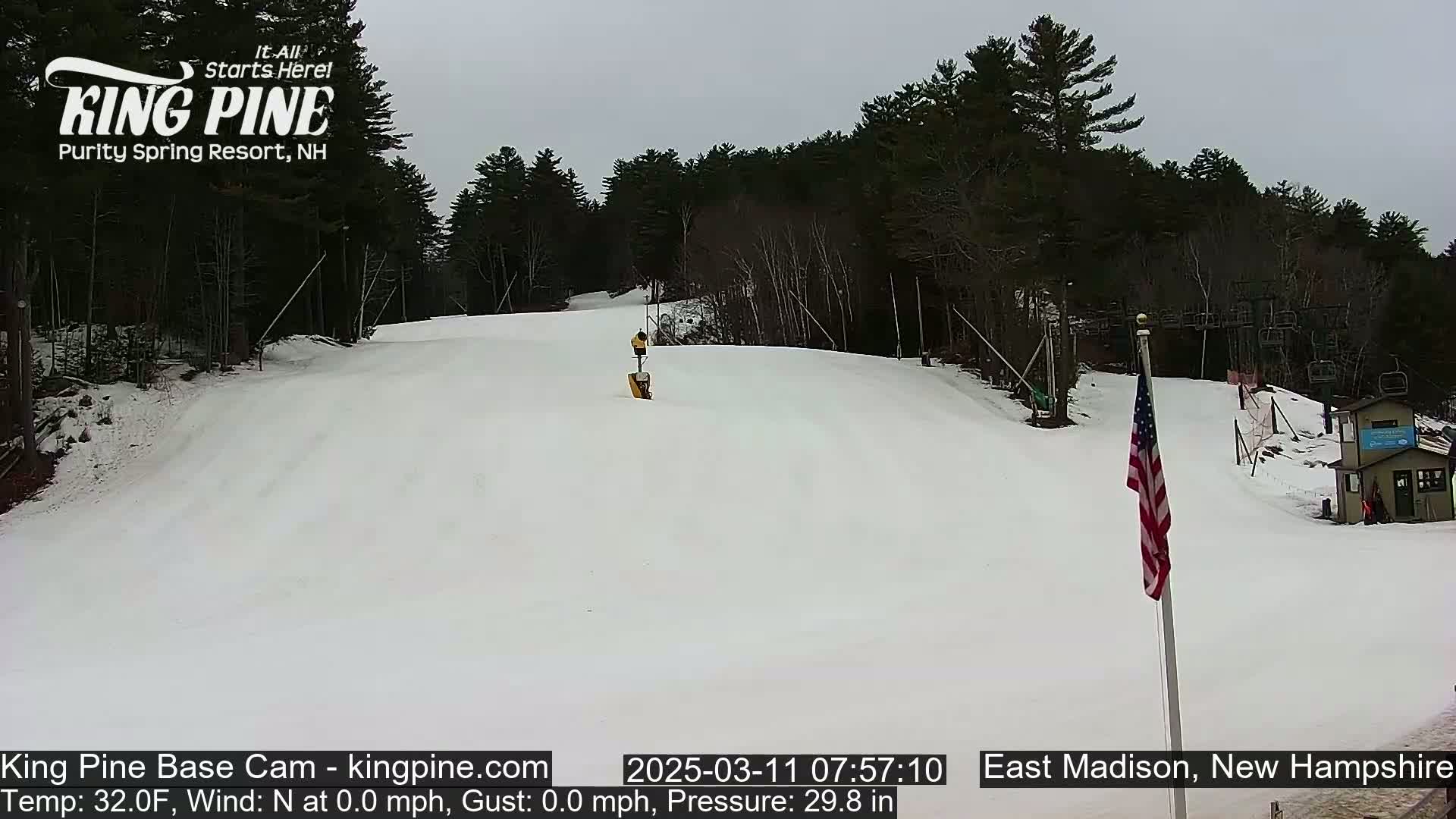 A snow-covered ski slope is visible, with a mostly cloudy sky overhead; a few skiers are on the slope and an American flag stands near a small building at the bottom.