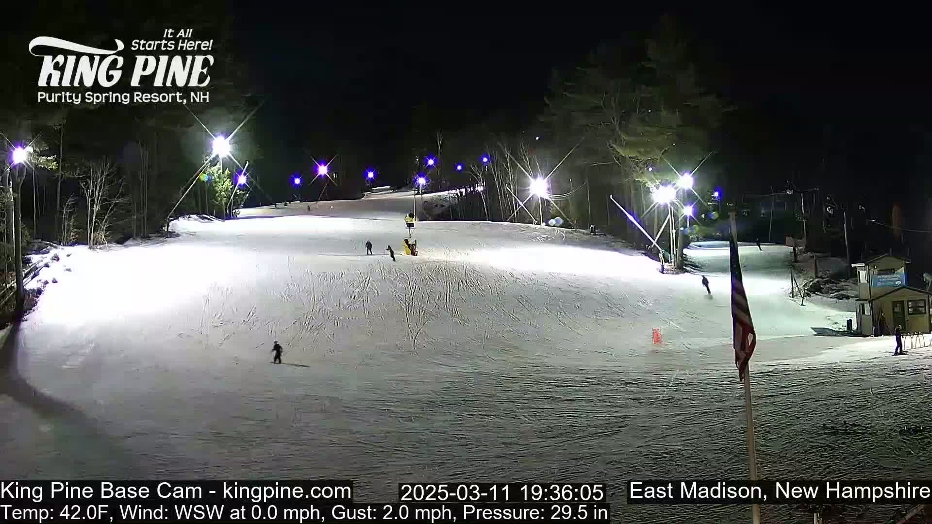 A snow-covered ski slope is illuminated at night by several bright lights, with a few skiers visible on the slope.