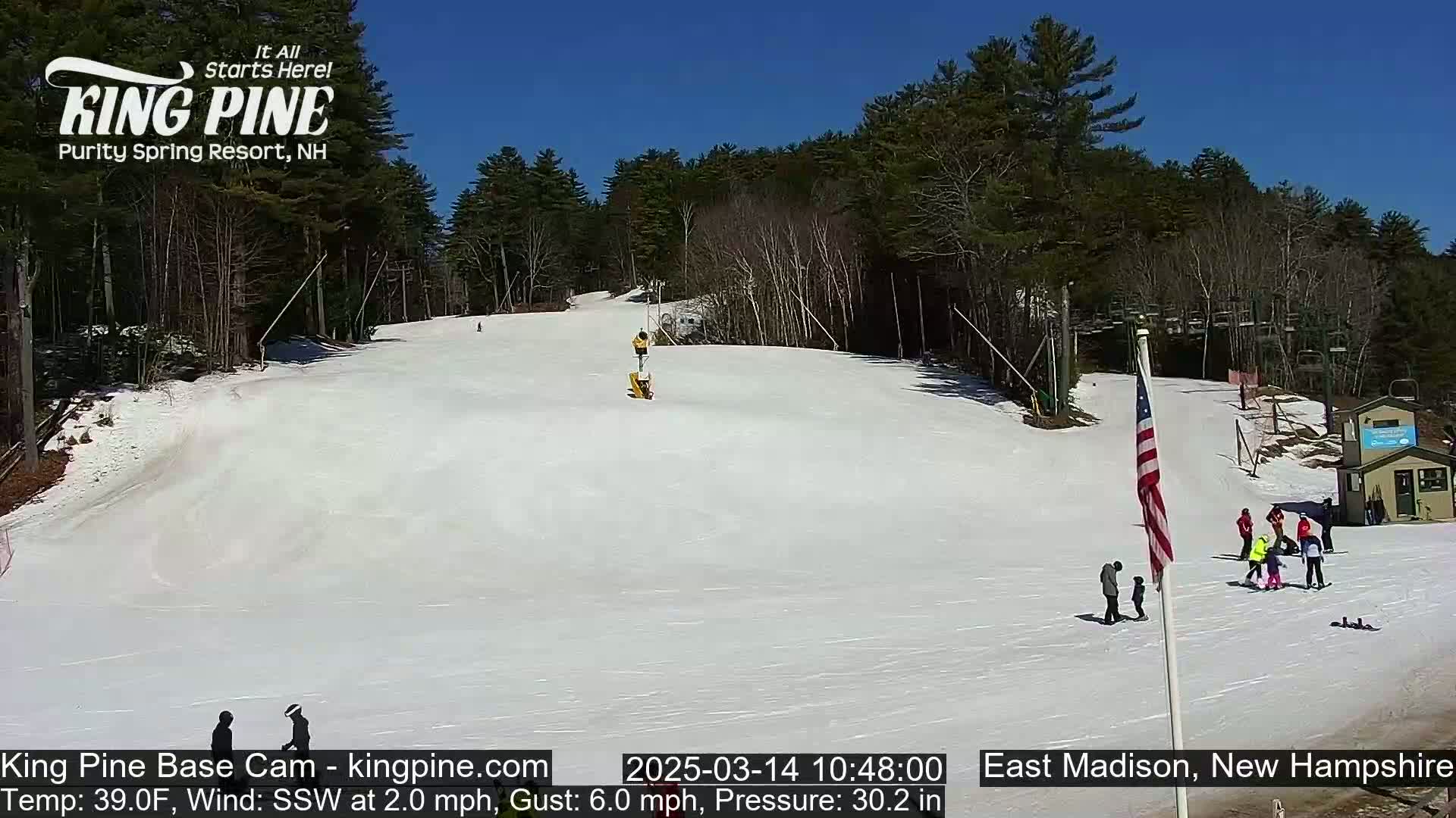 A snow-covered ski slope with several people at the bottom, under a clear blue sky.