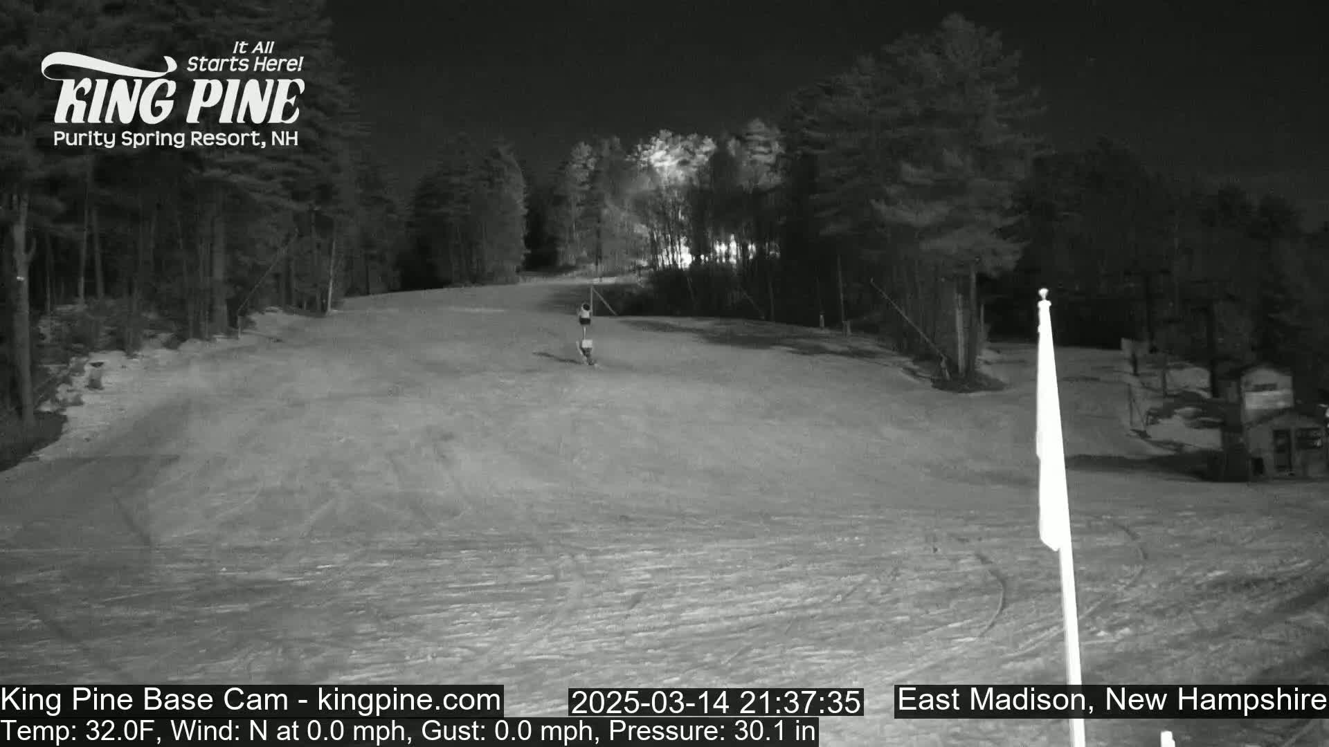 A nighttime, snow-covered ski slope is illuminated, with trees lining the sides and a single person visible in the distance.