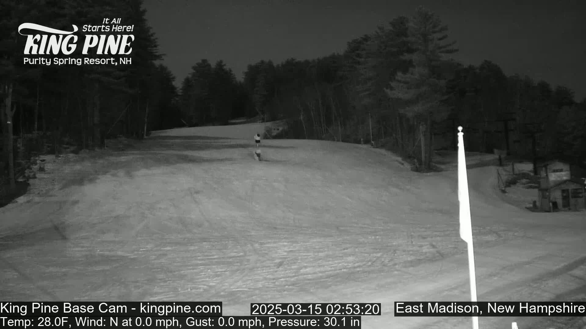 A snow-covered ski slope at night, with a single person visible near the bottom, under a dark sky.