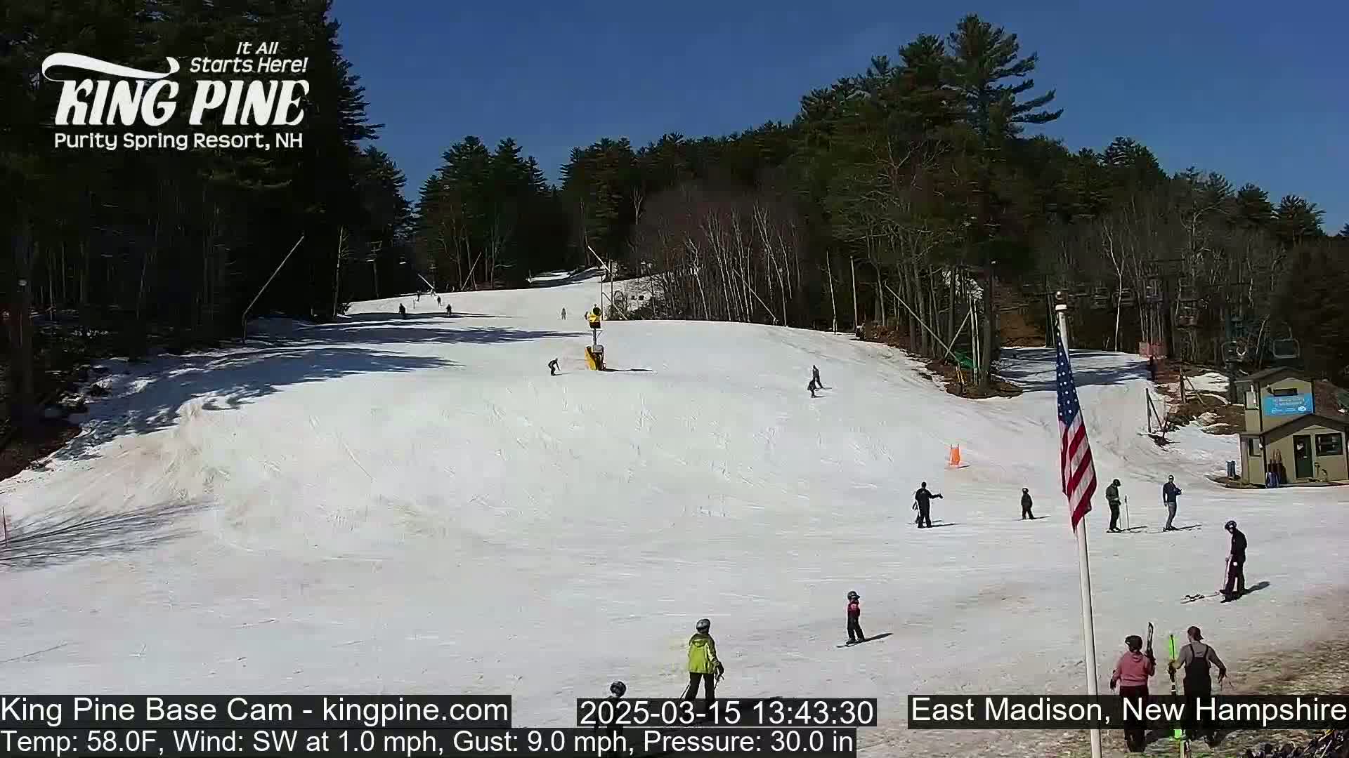 A sunny snow-covered ski slope with several skiers and snowboarders, surrounded by trees under a clear blue sky.