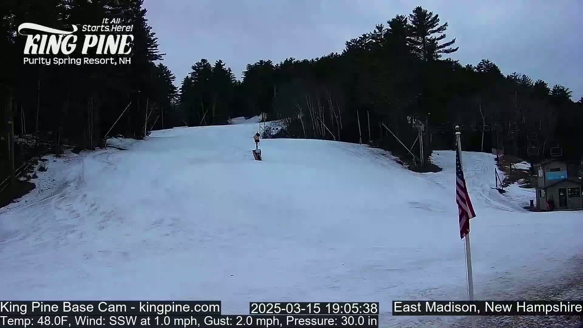 A snow-covered ski slope gently descends toward a small building, framed by a dark forest under a cloudy sky.