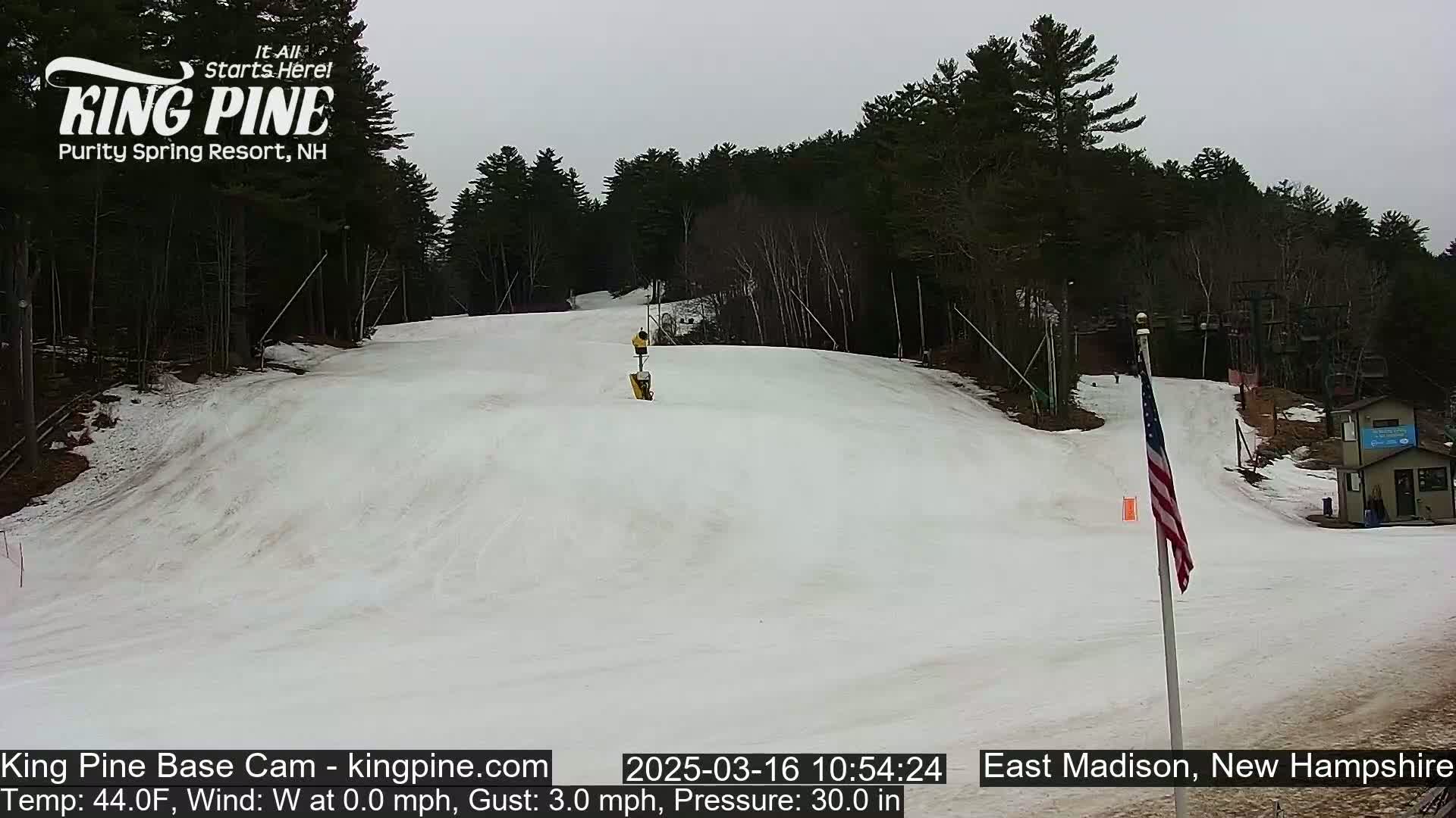 A snow-covered ski slope is visible under an overcast sky, with a sparsely populated wooded area in the background.