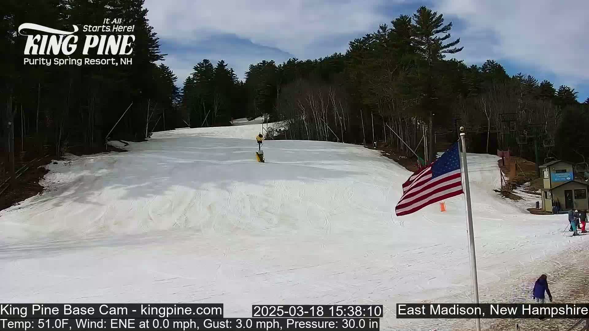 A snow-covered ski slope with a few people visible, under partly cloudy skies, is framed by a wooded area with an American flag in the foreground.