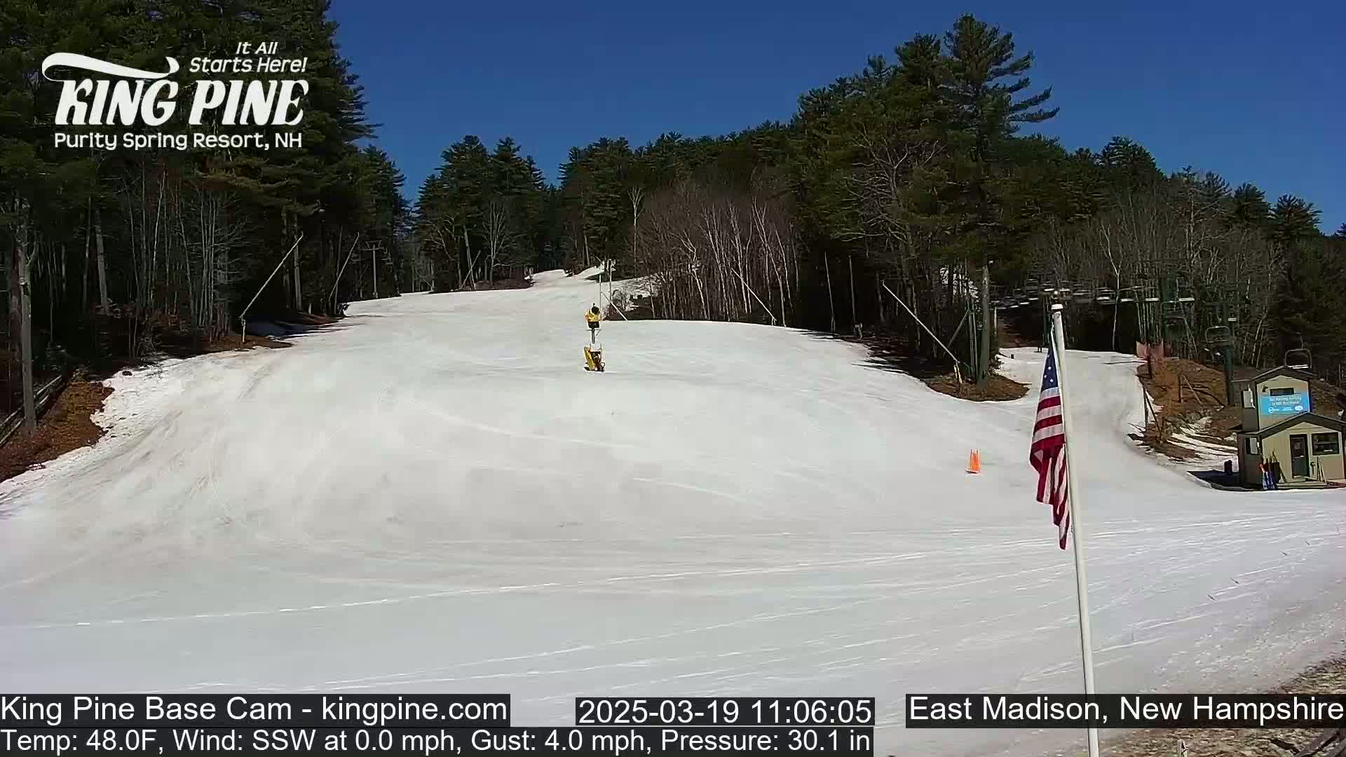 A mostly sunny day shows a snow-covered ski slope with a person skiing down, lift towers, and an American flag; trees line the background.