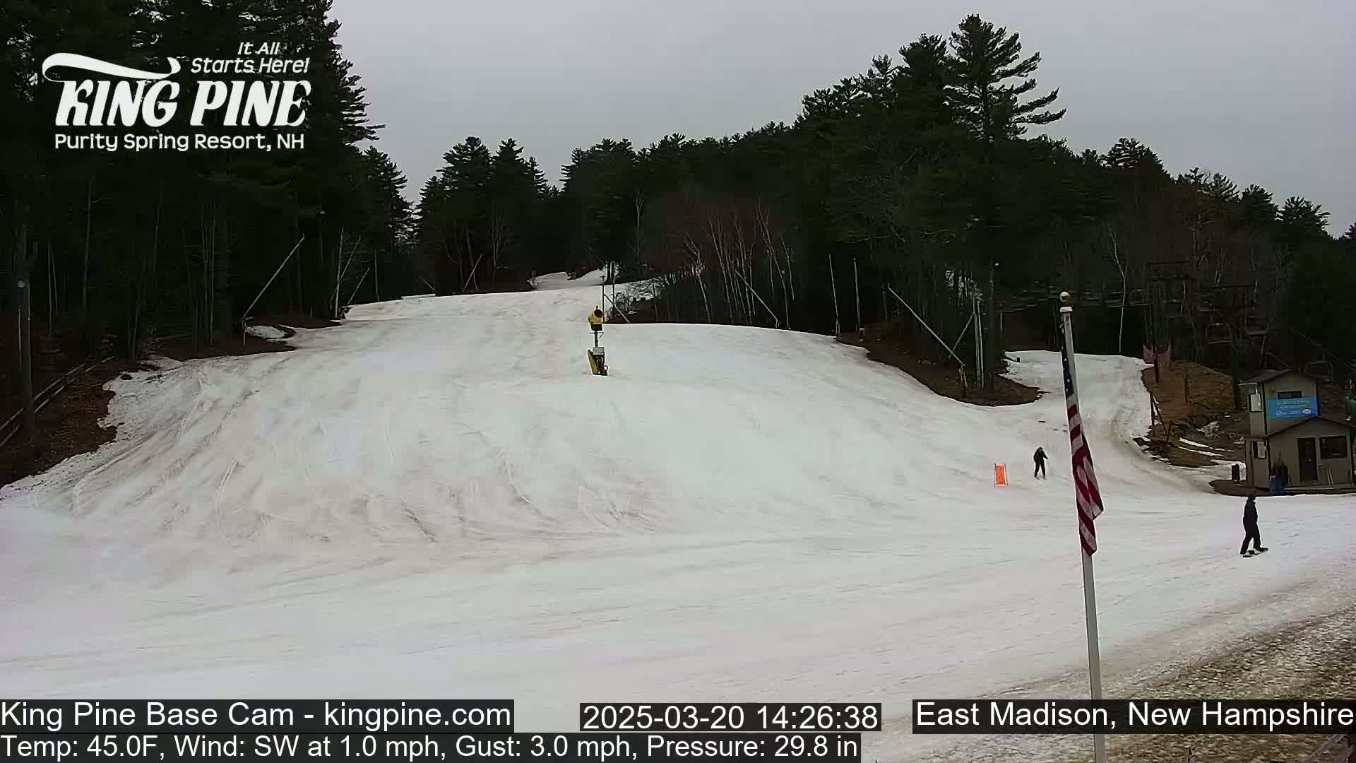 A snow-covered ski slope with a few people skiing or snowboarding, under an overcast sky.