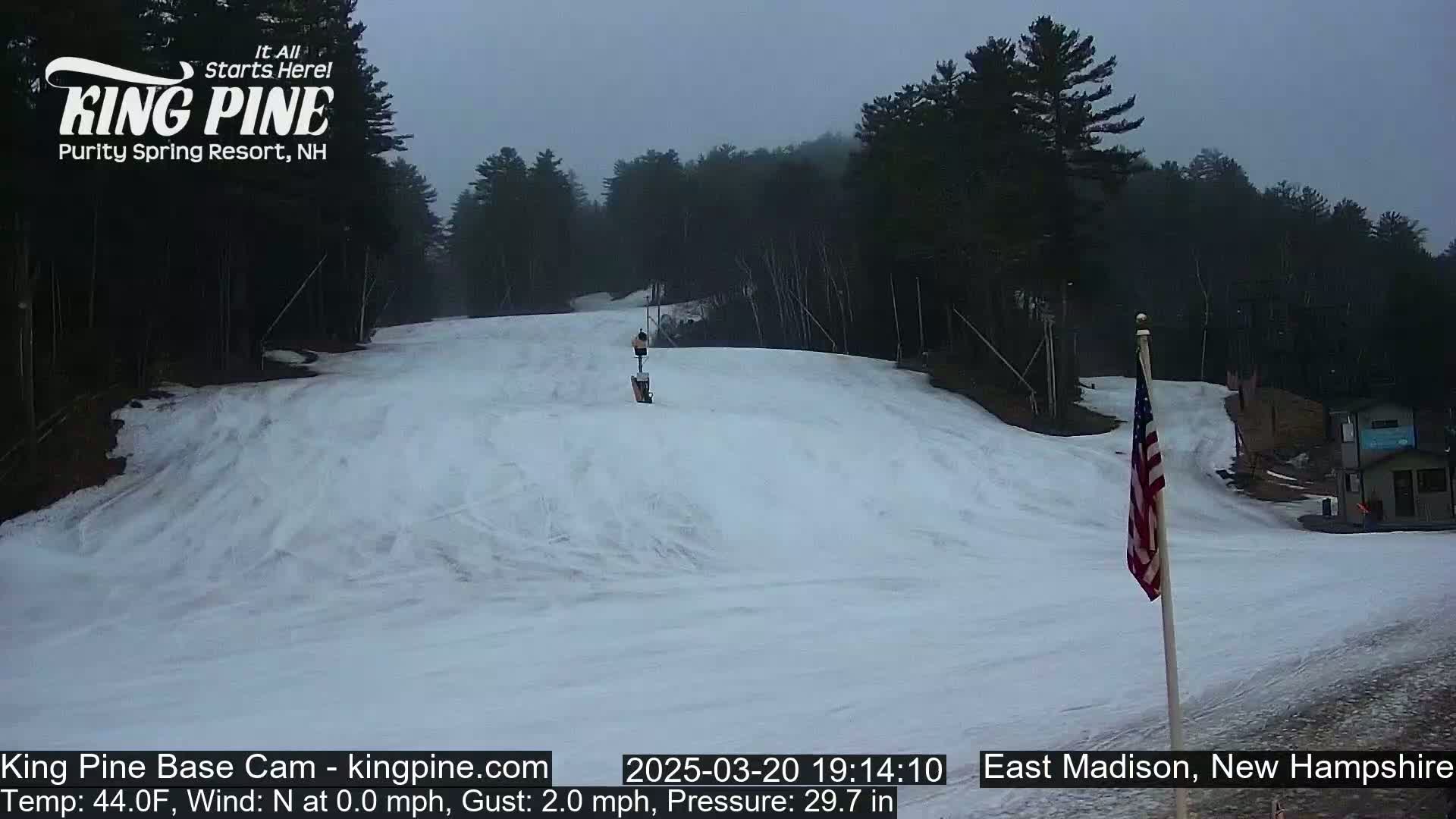 A snow-covered ski slope is visible under an overcast sky, with a lone person skiing down the hill and an American flag standing in the lower right corner.