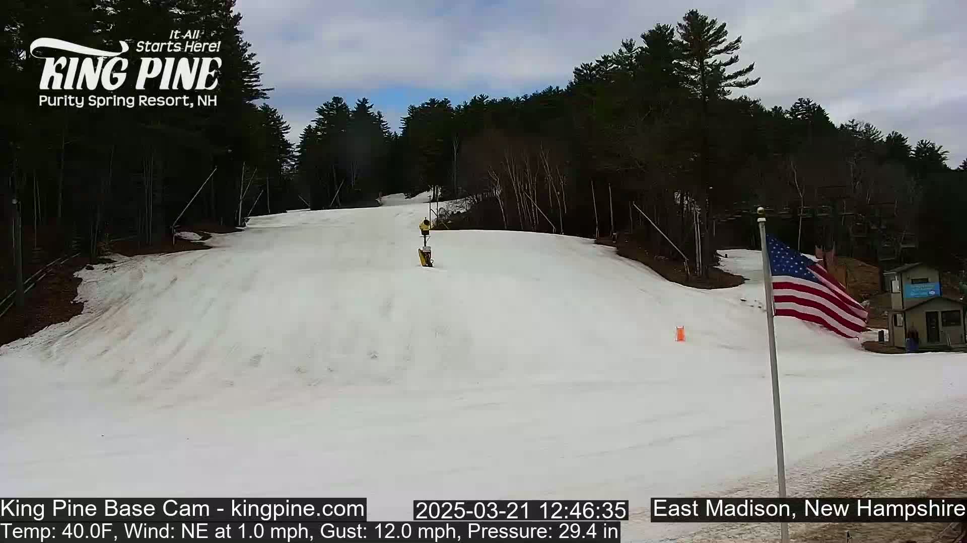 A snow-covered ski slope is visible beneath a mostly cloudy sky, with a person skiing down the hill and an American flag waving gently in the background.