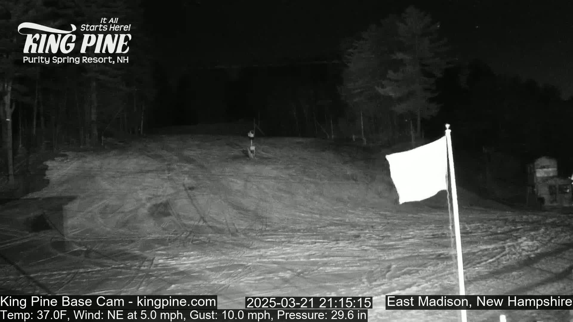 A nighttime, snow-covered slope with a white flag on a pole, under a dark sky with a northeast wind.