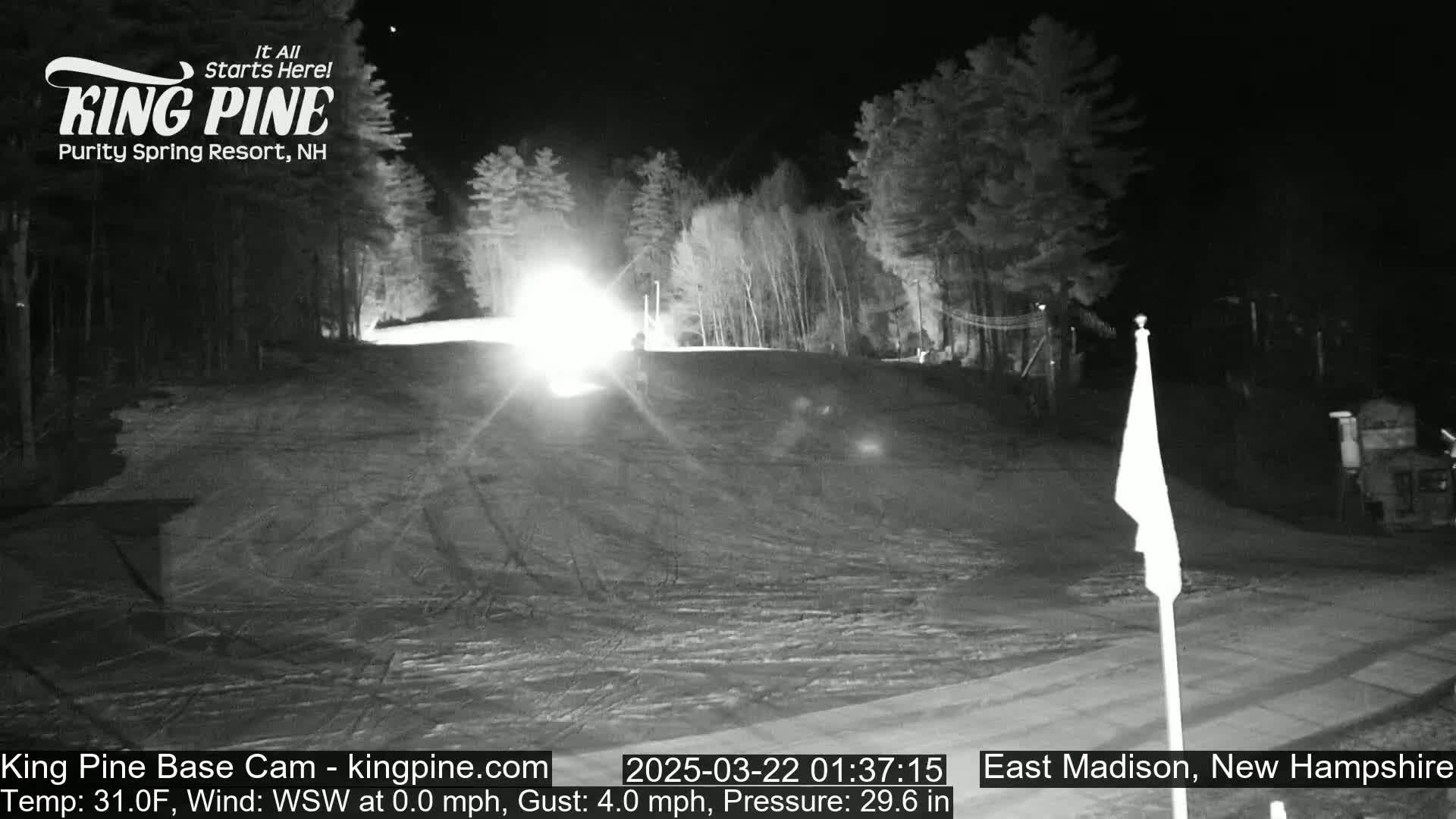 A nighttime, snow-covered ski slope is illuminated by a bright light, with trees lining the sides under a clear sky.