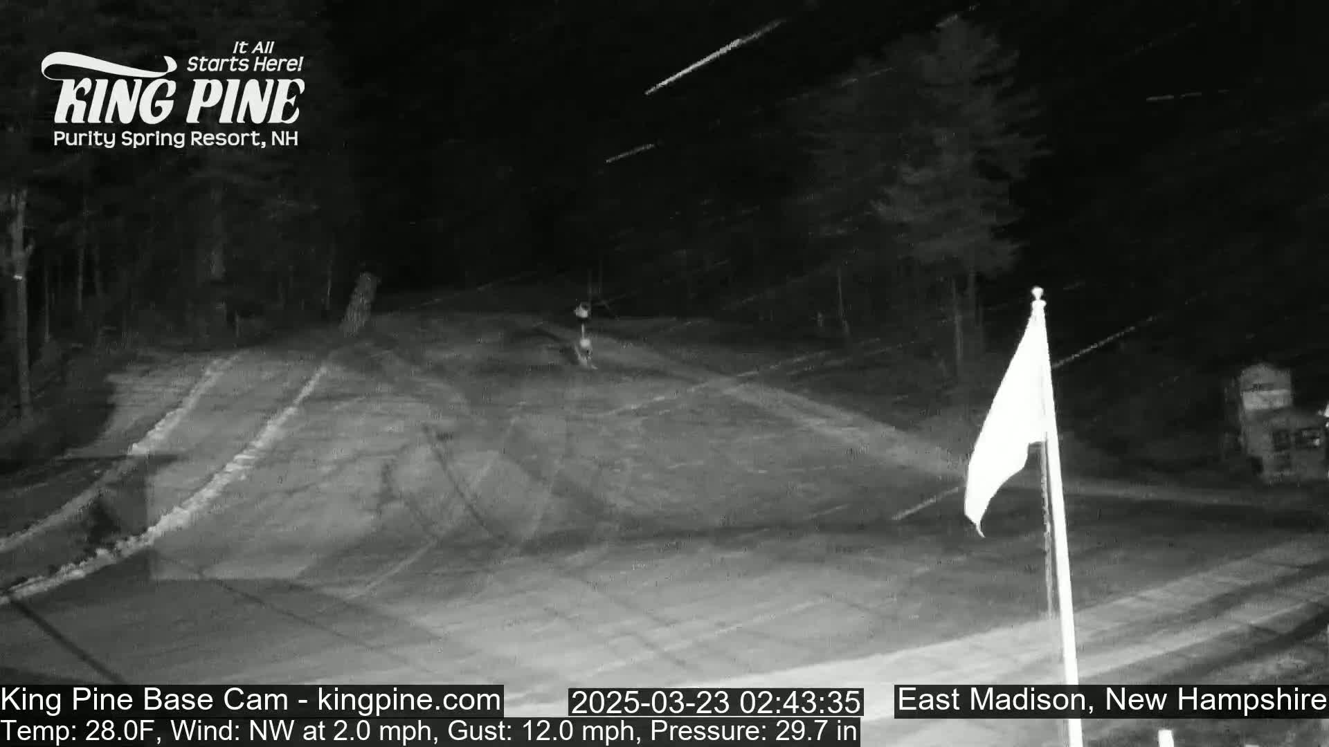 A nighttime, snowy scene shows a mostly empty paved area with a flagpole and trees in the background.