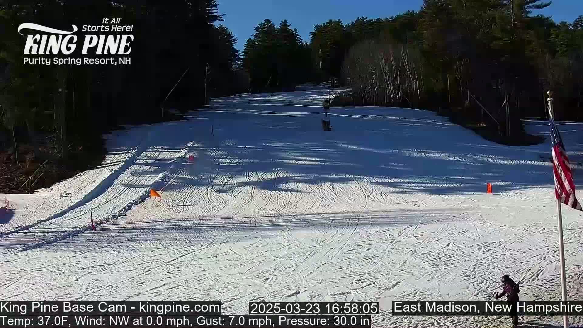A snow-covered ski slope, under a clear blue sky, is bordered by dark evergreens.