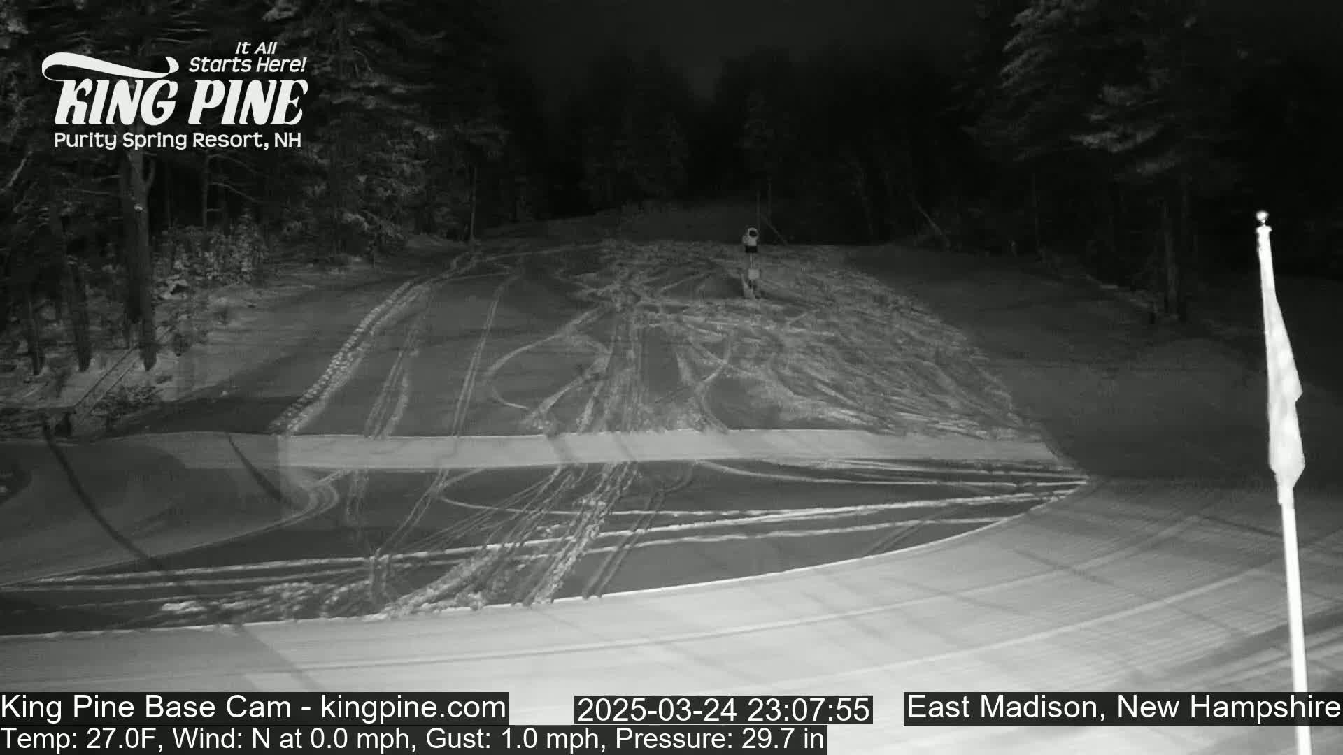 A snow-covered ski slope at night, illuminated under dark skies with faint tracks in the snow.