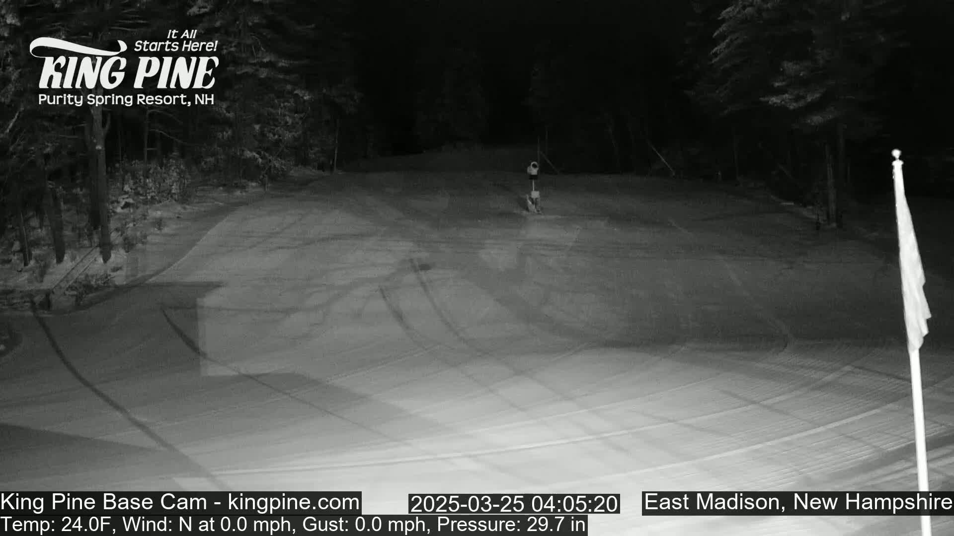 A snow-covered, nighttime scene shows a mostly empty ski slope with trees in the background under a dark sky.