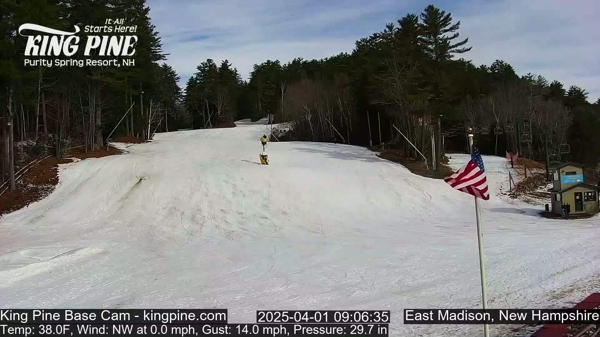 A snow-covered ski slope with a few skiers, a chairlift, and an American flag, under a partly sunny sky.