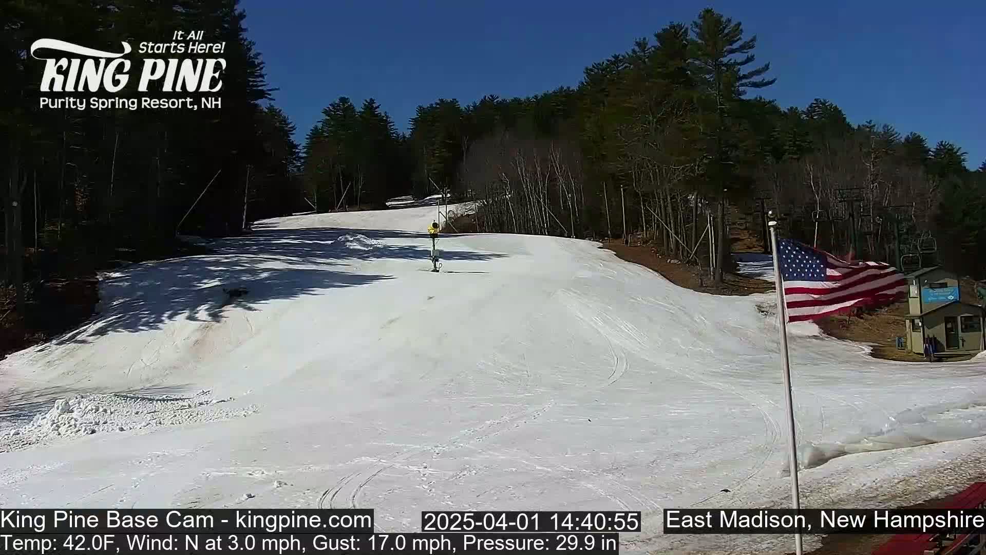 A snow-covered ski slope is visible under a clear blue sky, with trees lining the background and a small American flag waving in the breeze.
