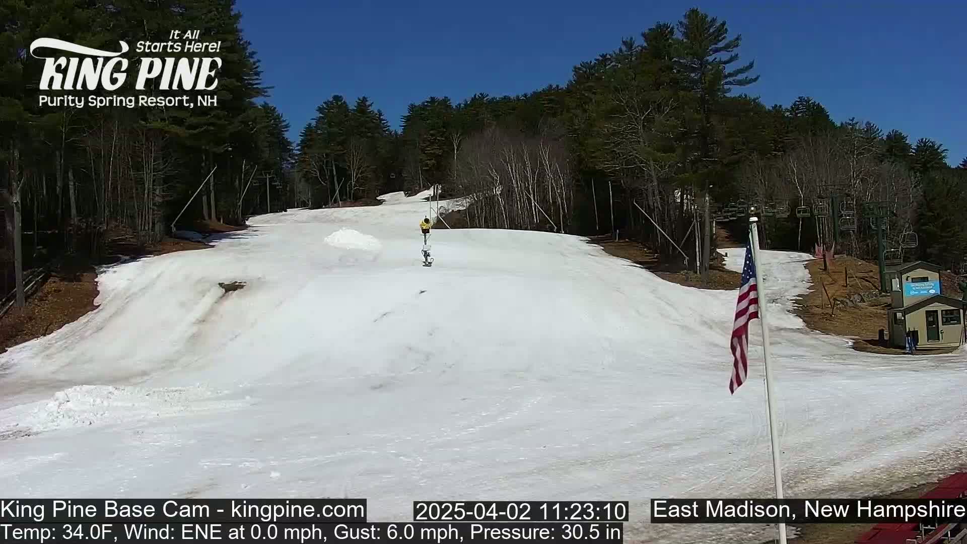 A person snowboards down a snow-covered slope on a sunny day, with a ski lift and trees visible in the background.