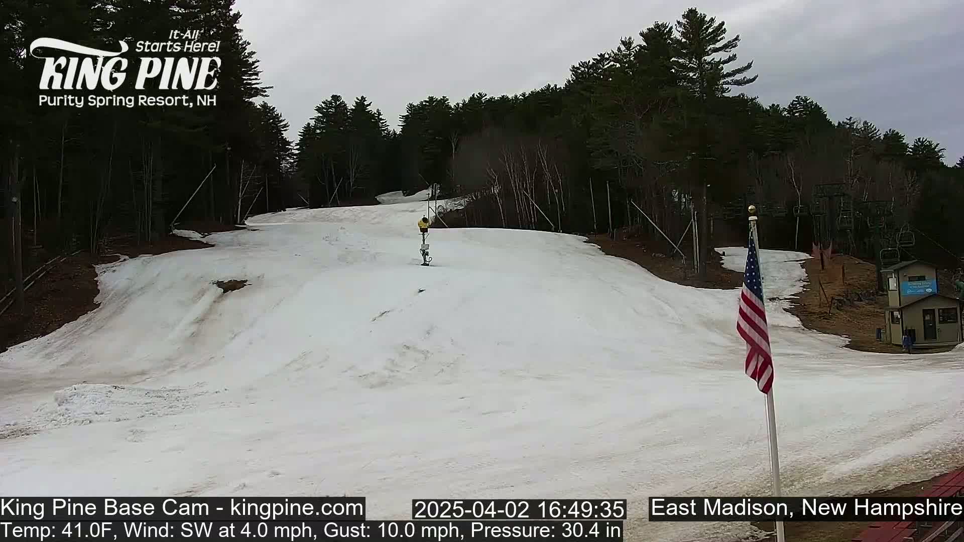 A snow-covered ski slope with a person skiing down it, under an overcast sky, is surrounded by trees.