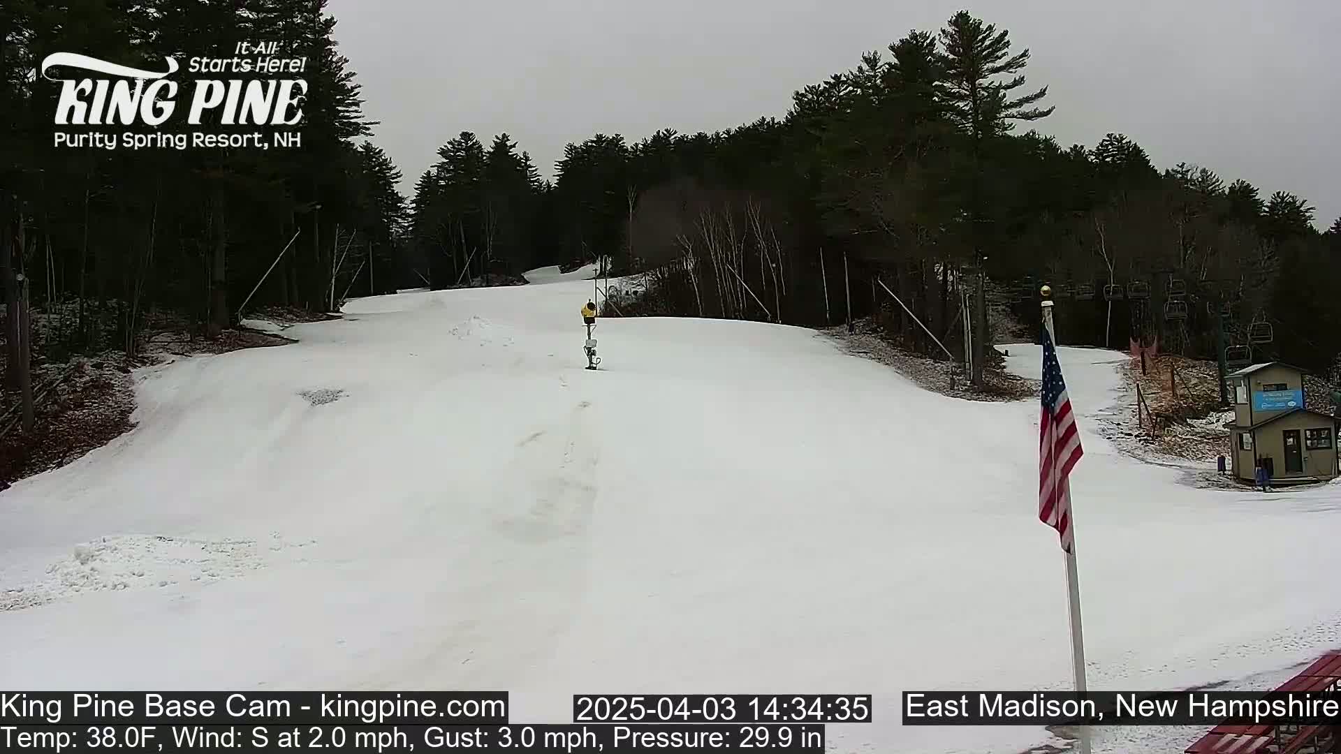 A person skis down a snow-covered hill in overcast weather, with a wooded area in the background and an American flag visible to the right.