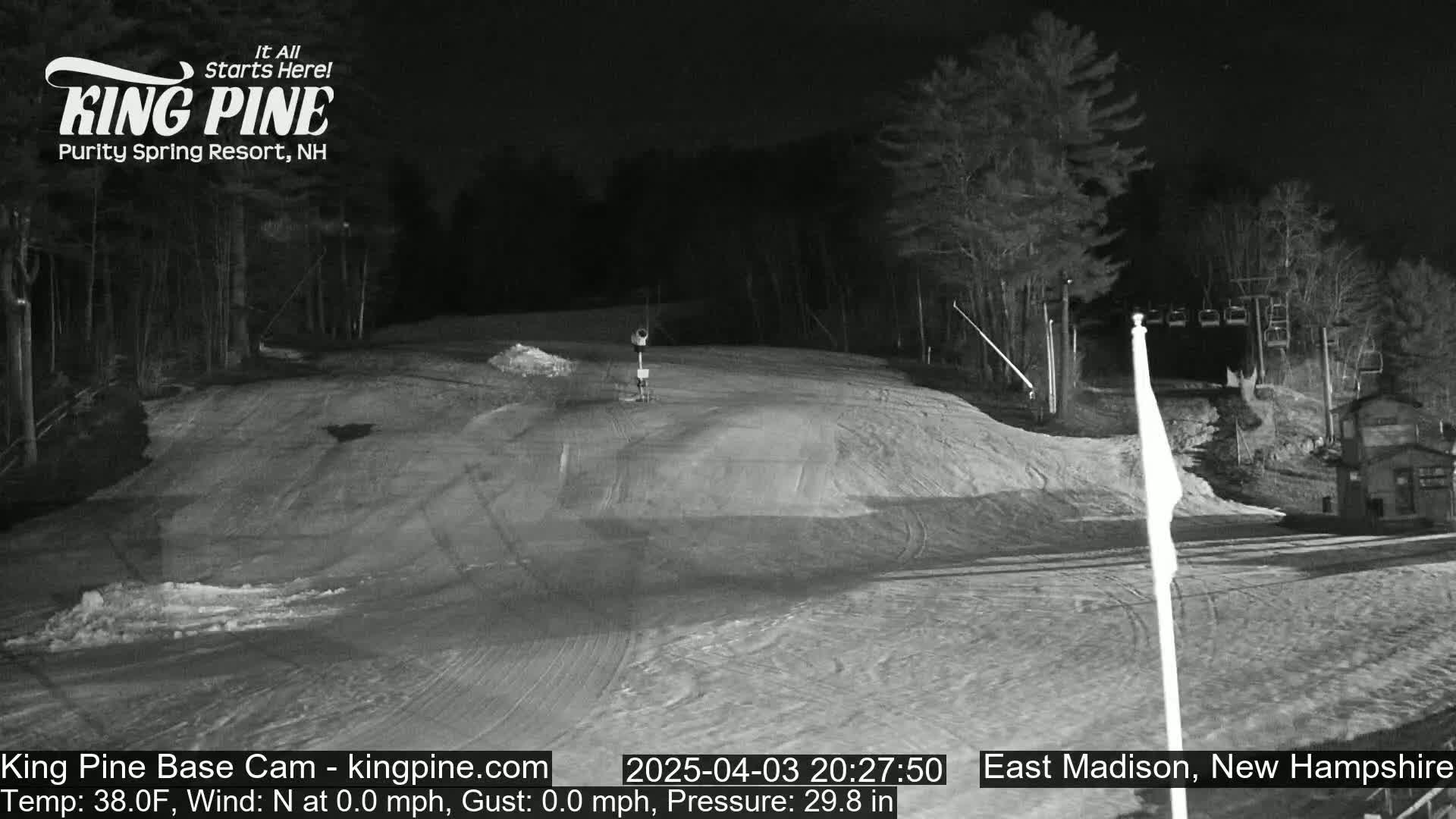 A nighttime, snow-covered ski slope is illuminated, showing a mostly empty run with a chairlift and some trees in the background.
