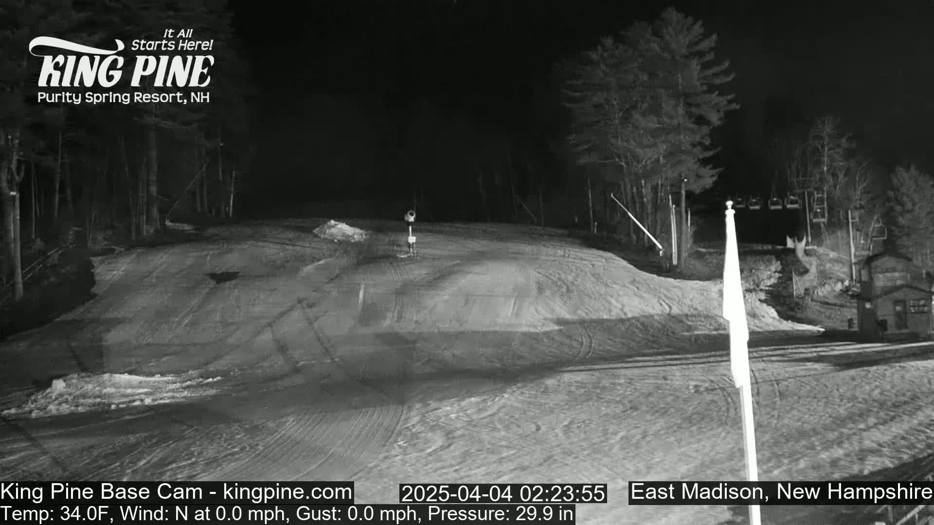 A snow-covered ski slope at night, illuminated by artificial light, with trees and a ski lift in the background.