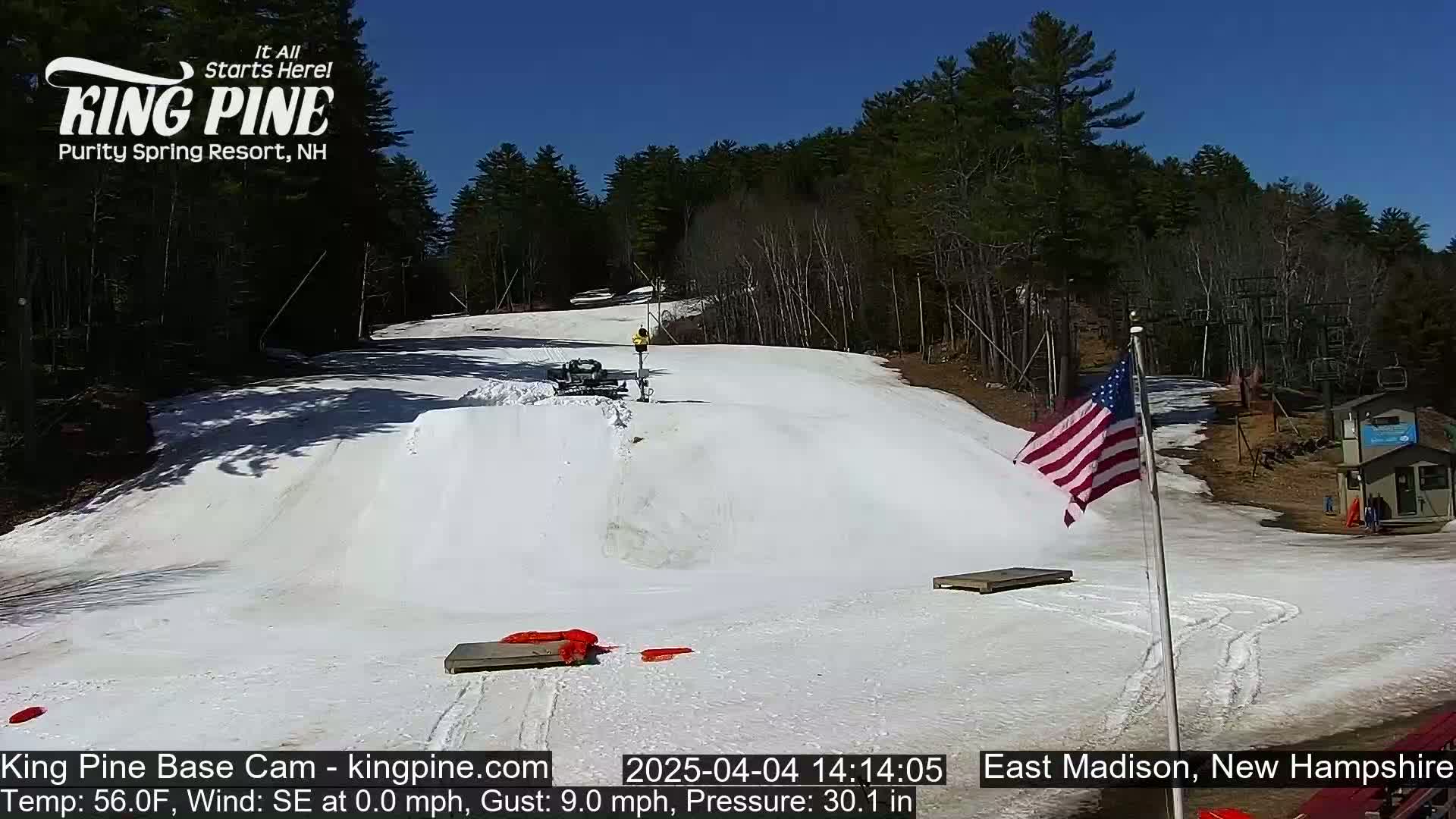 A snow-covered ski slope with a snow groomer and a person visible, under a clear blue sky, is situated next to a wooded area with a small American flag waving in the wind.