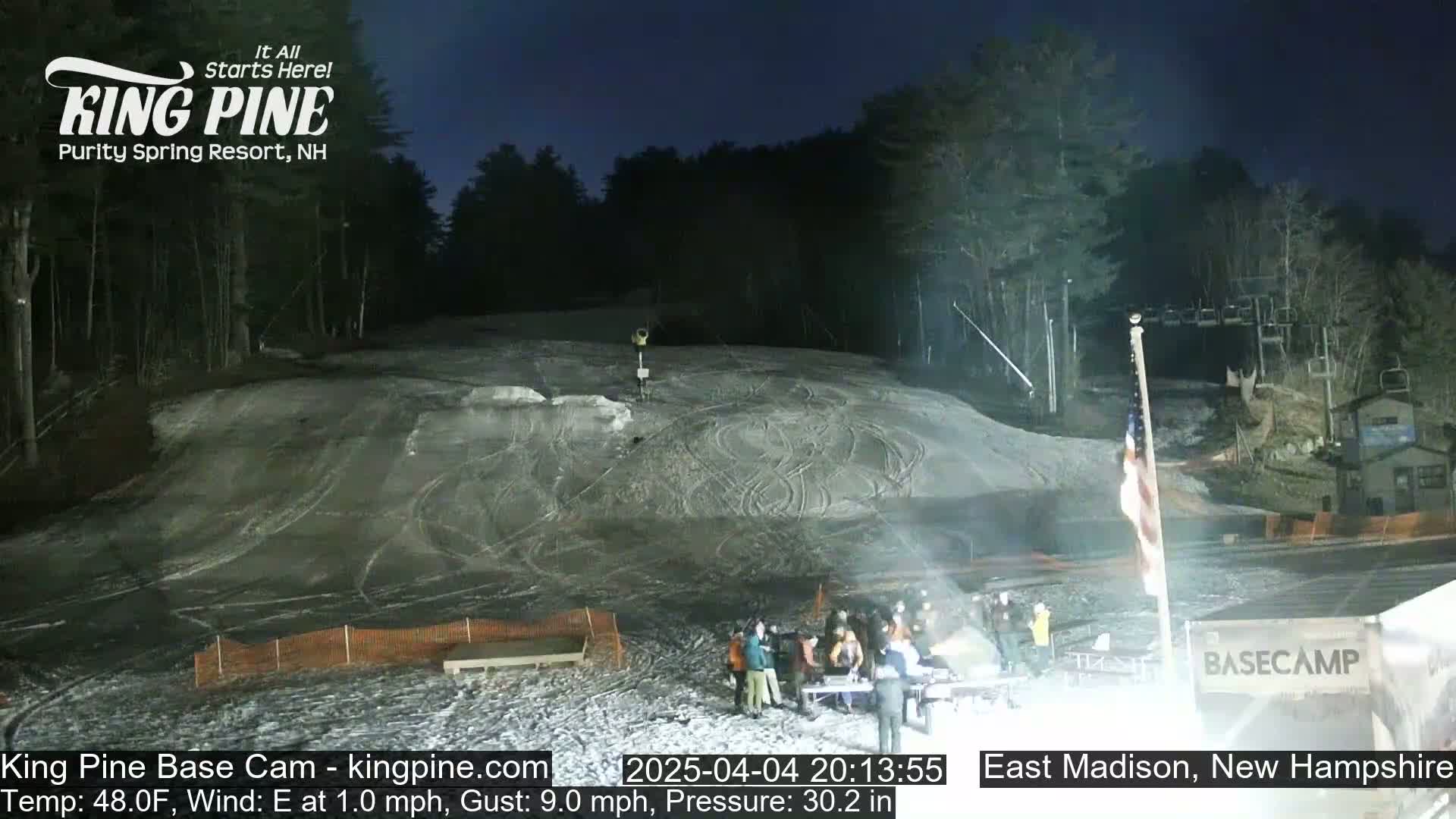 A snow-covered ski slope at night, illuminated by artificial lights, with a group of people gathered near a basecamp building under a dark sky.