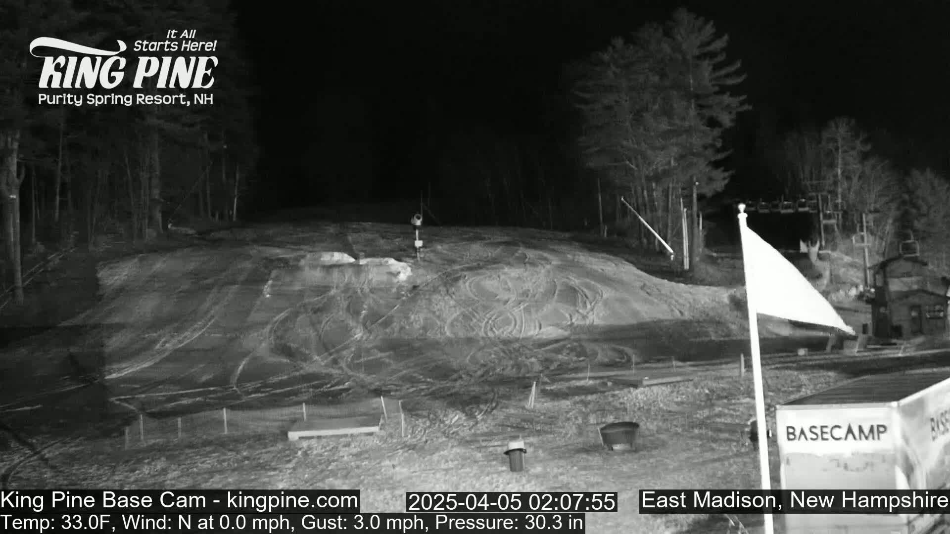 A nighttime, snow-covered ski slope with a few structures and trees in the background under a clear sky.