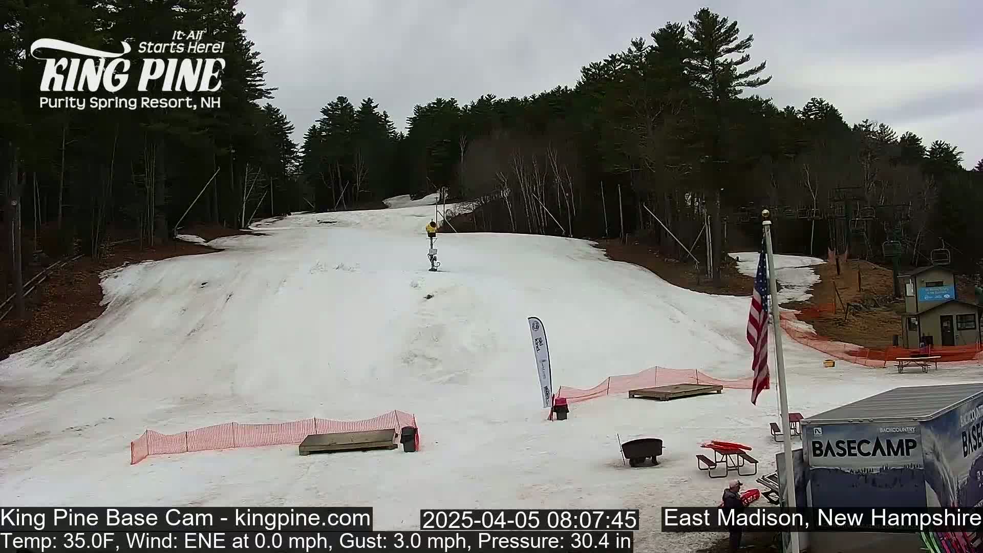 A snow-covered ski slope with a few people on it, under an overcast sky, is surrounded by trees and has some picnic tables and other equipment at the bottom.
