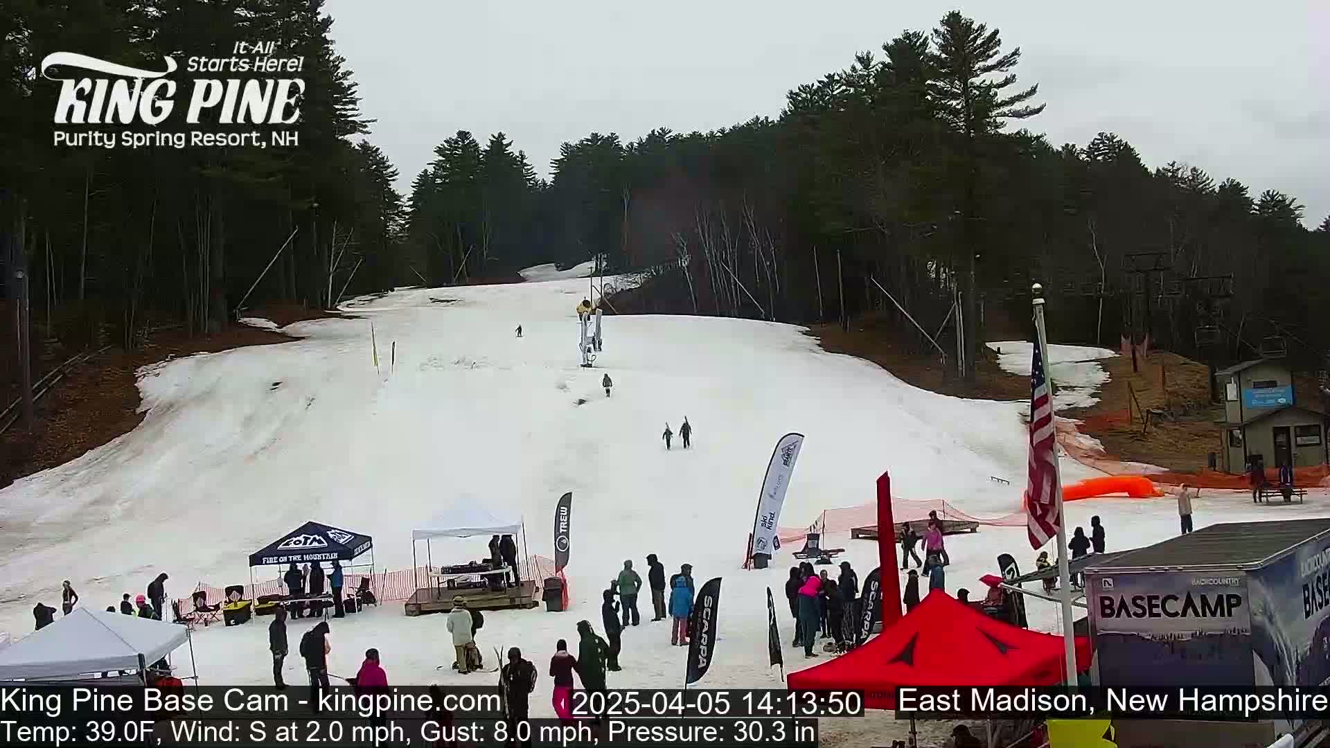 A snow-covered ski slope with several people skiing and snowboarding, along with spectators and tents at the base of the hill, under an overcast sky.