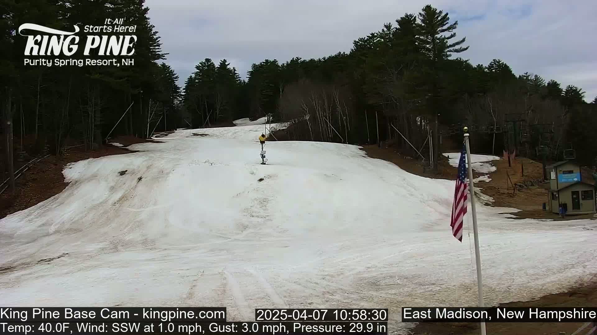 A snow-covered ski slope with a person skiing down it, under a cloudy sky, is surrounded by trees.