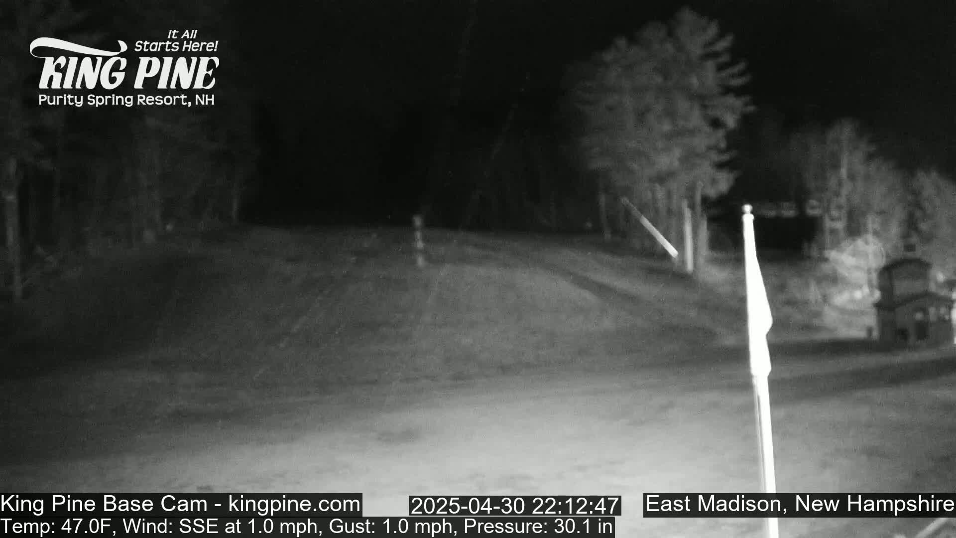 A nighttime, low-light outdoor scene shows a road curving through a wooded area, with a flagpole visible in the foreground, under a clear sky and with a temperature of 47°F and a gentle SSE wind.