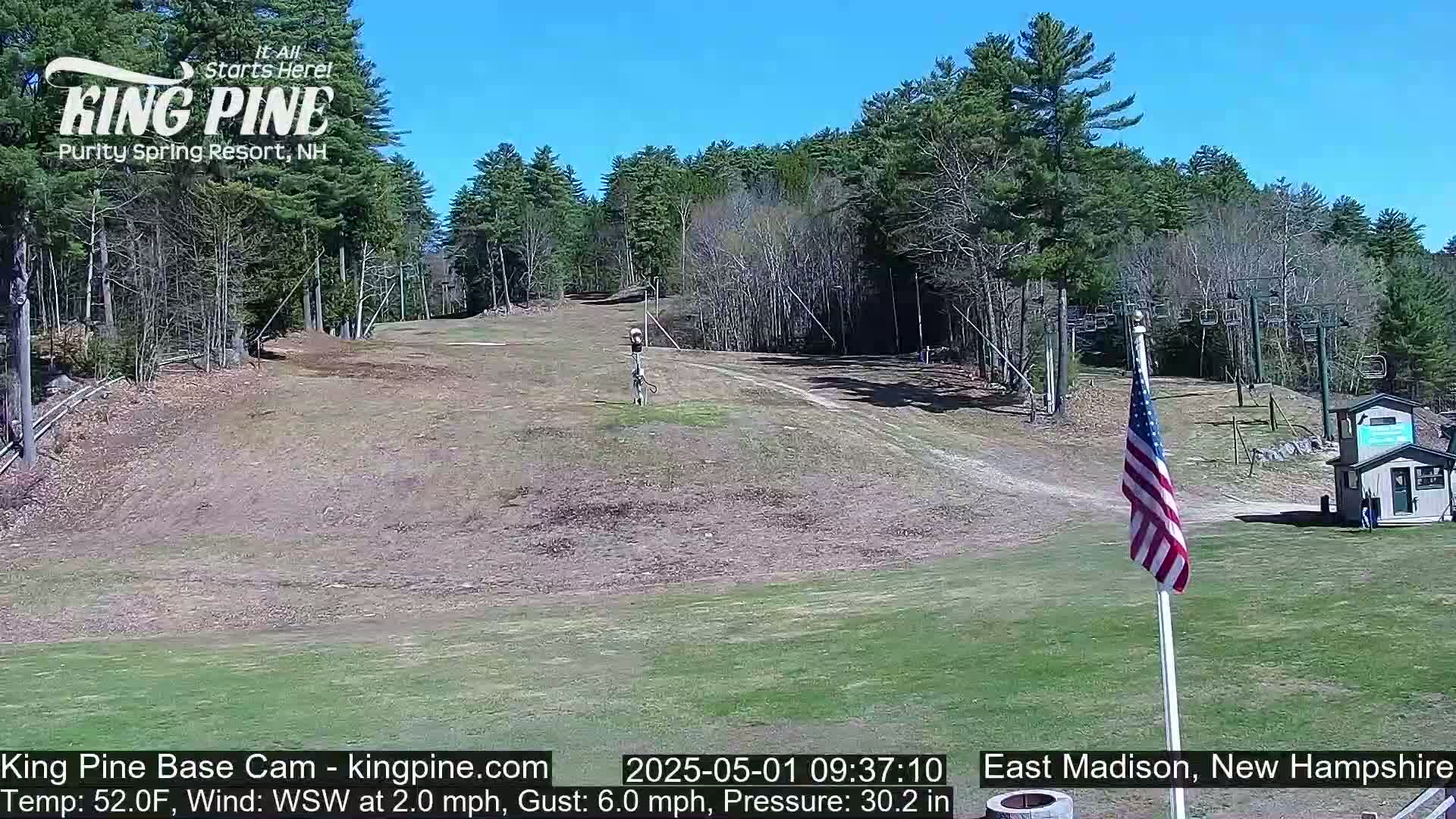 A ski slope with a chairlift and an American flag is visible on a sunny day, surrounded by a forest of deciduous trees.