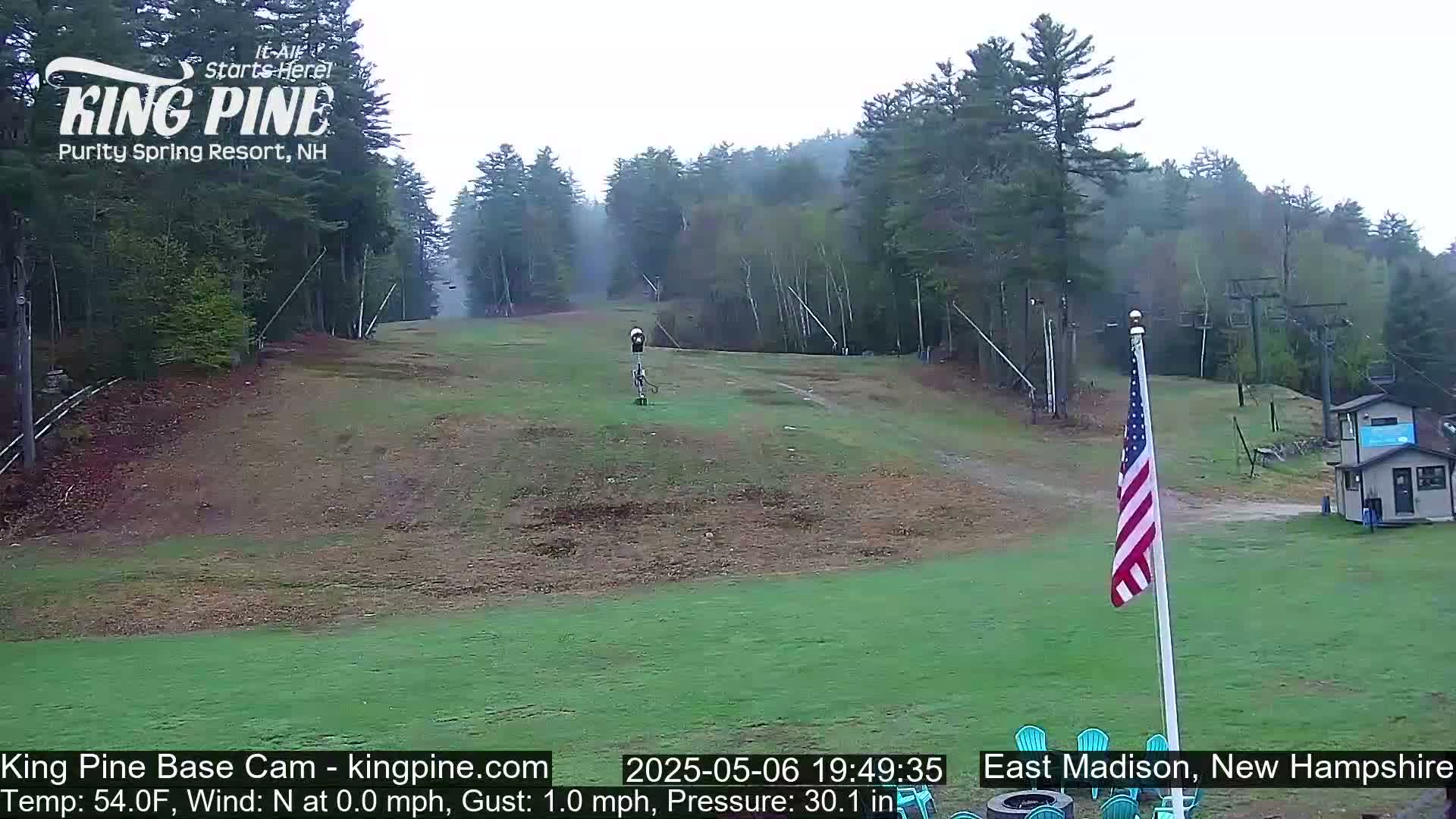 A mostly clear view of a ski slope in the daytime, with a small building and an American flag visible in the foreground, and a light wind.