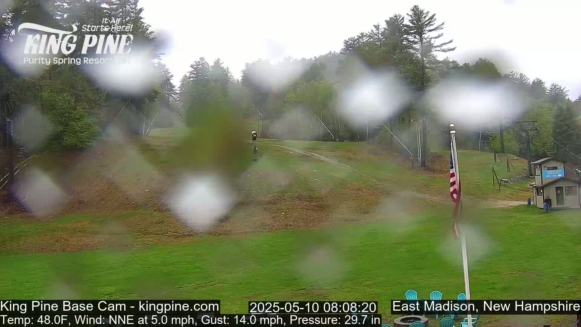 A rainy view of a grassy area with a ski lift and a small building in the background, surrounded by trees.