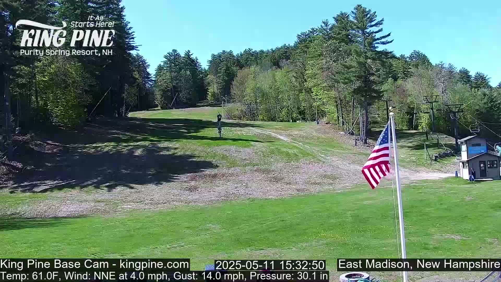 A grassy ski slope is visible under a clear blue sky, with a forested hillside in the background and an American flag waving near a small building.