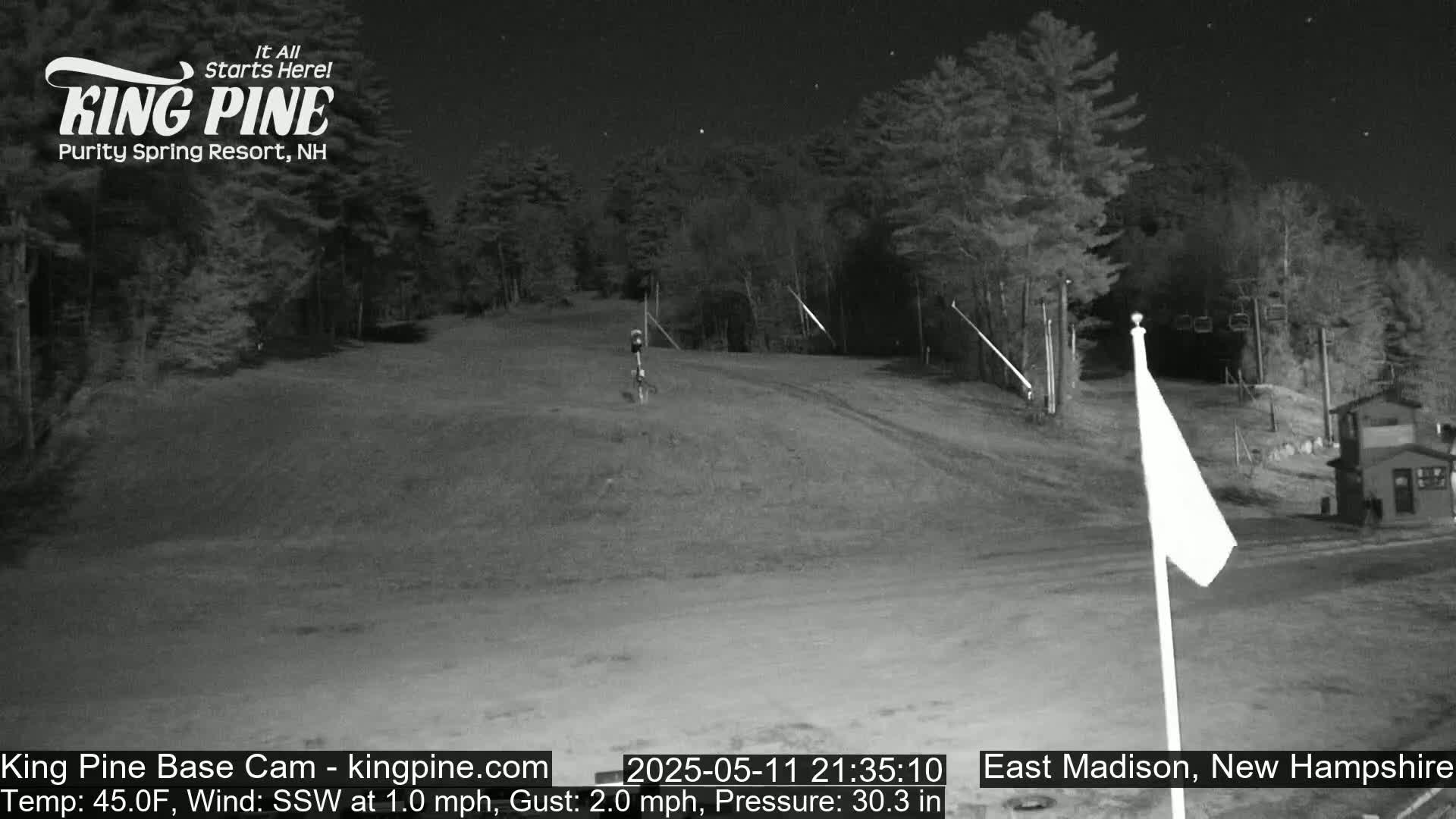 A nighttime view of a ski slope under a clear, starry sky, with trees lining the sides and a flagpole in the foreground.