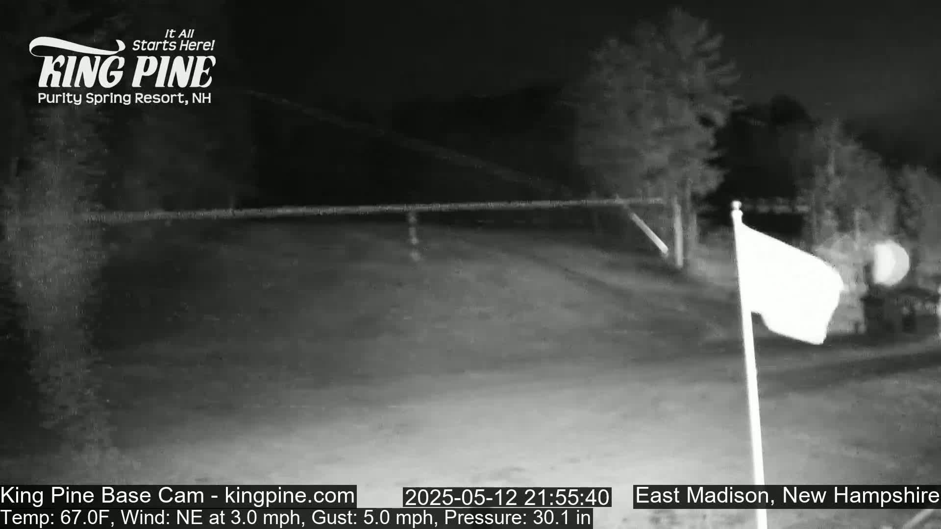 A nighttime, low-light view of a mostly empty paved area with a flagpole and a white flag blowing gently in a northeast wind.