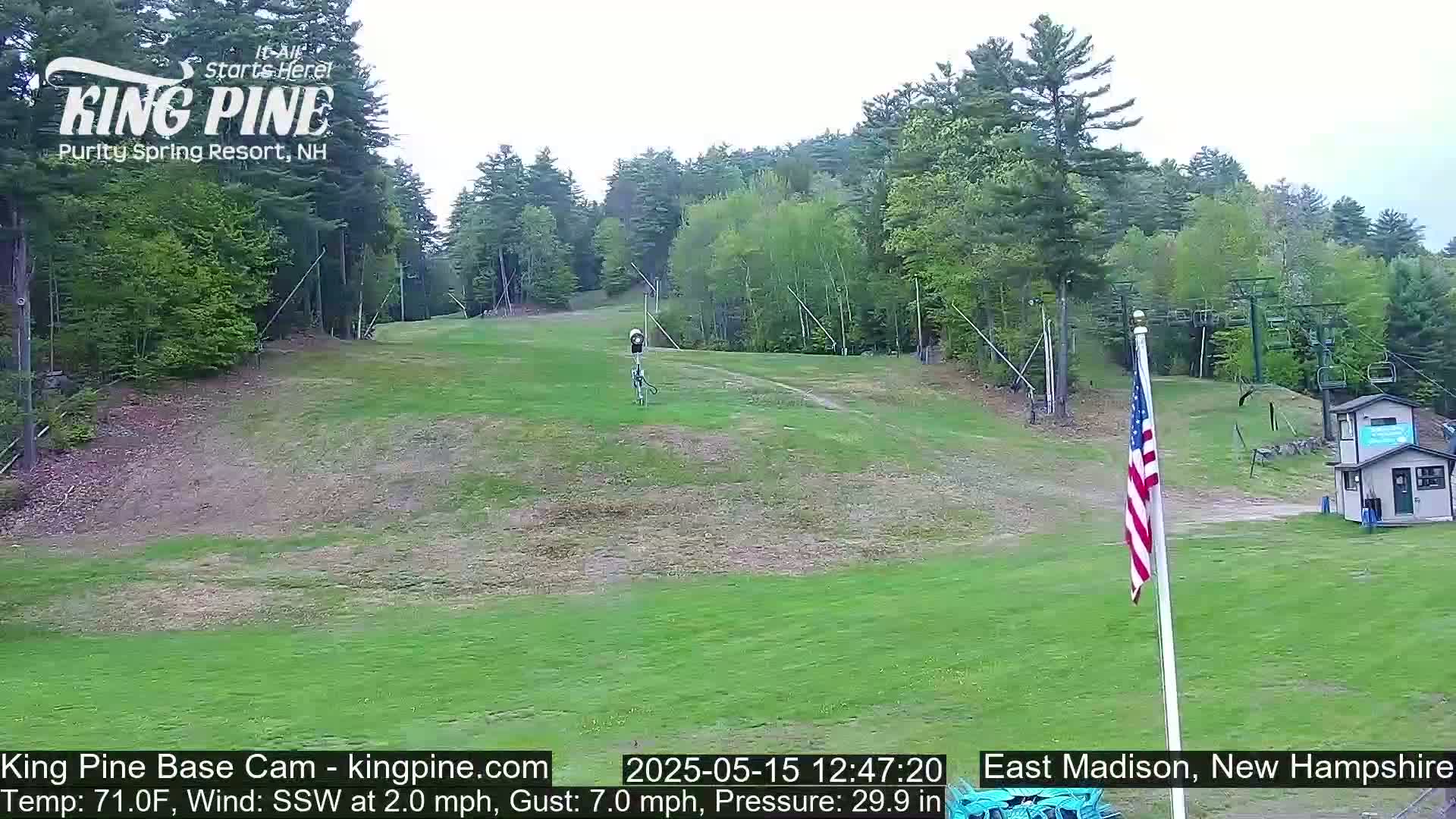 A mostly sunny view of a grassy ski slope with a chairlift, a small building, and an American flag, under partly cloudy skies.
