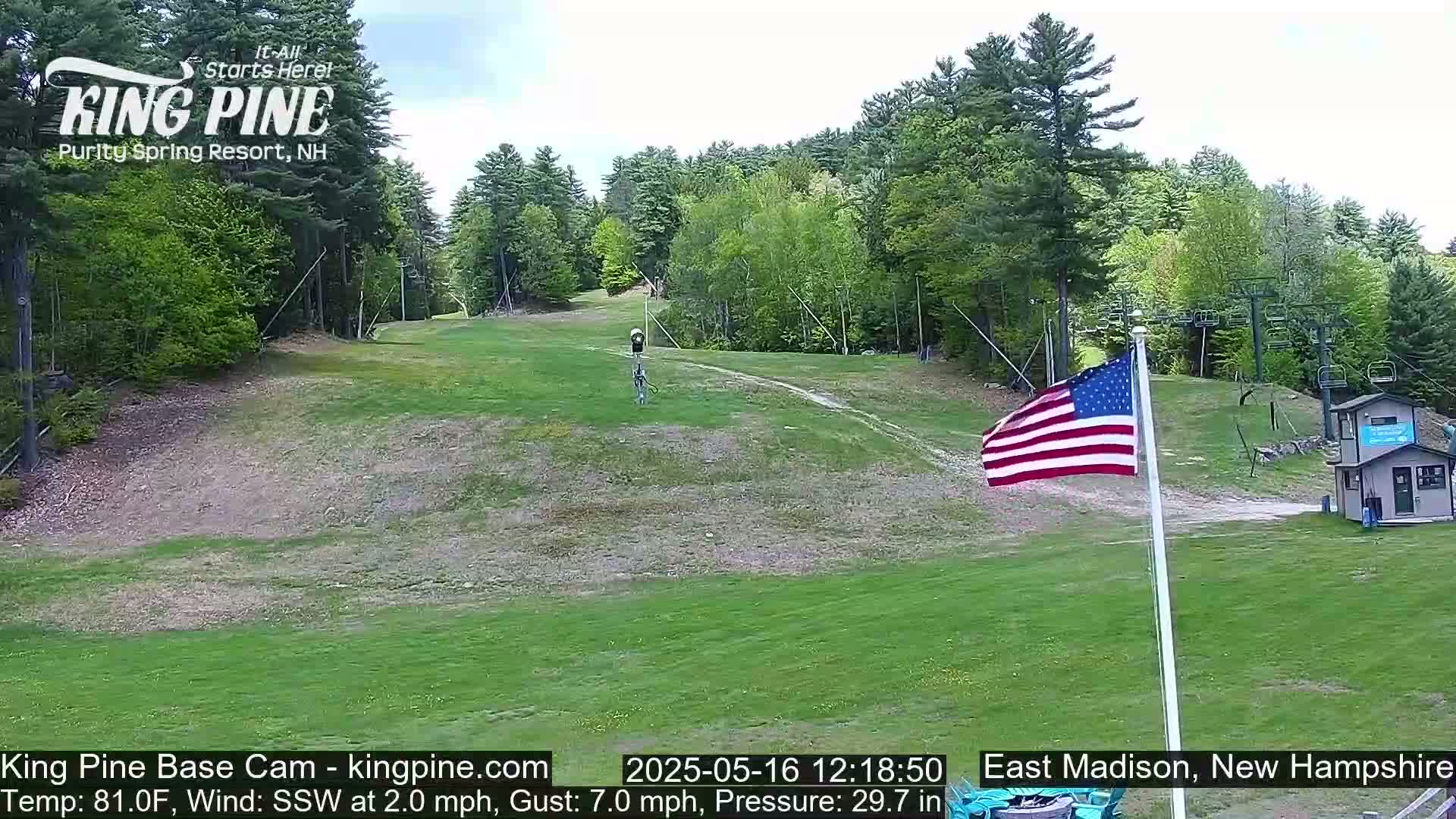 A grassy ski slope with a chairlift, a small building, and an American flag waving in a light south-southwesterly breeze on a partly sunny day.