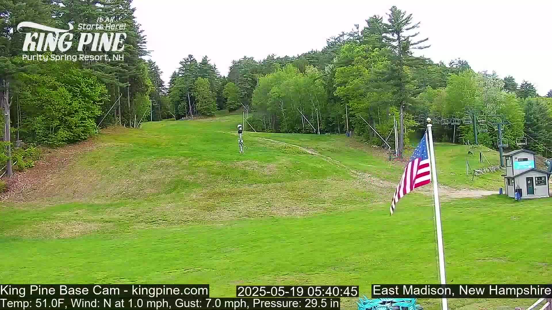 A grassy ski slope with a chairlift and American flag is visible under a partly cloudy sky.