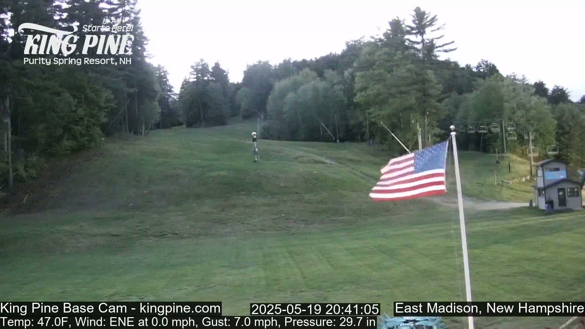 A grassy hill with a ski lift in the background under a partly cloudy sky, with an American flag waving in the foreground.
