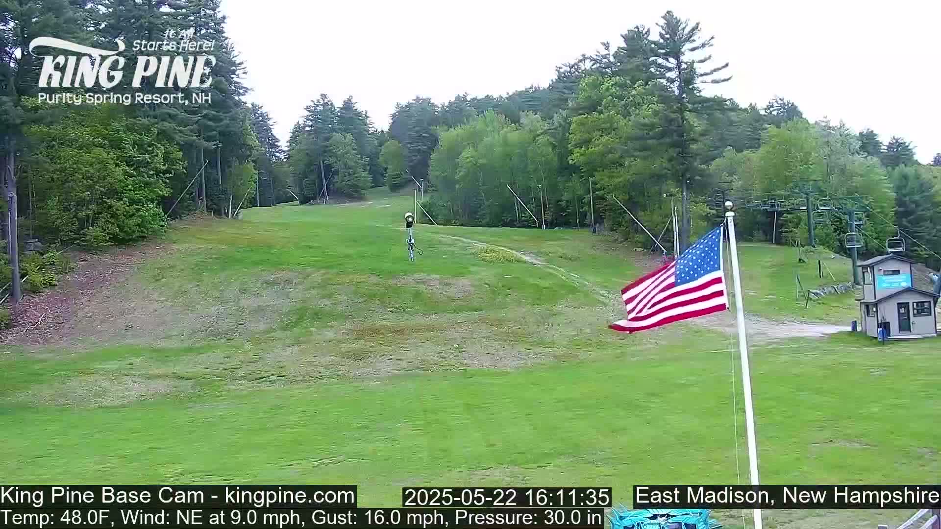 A grassy ski slope with a ski lift and an American flag in the foreground, under a cloudy sky.