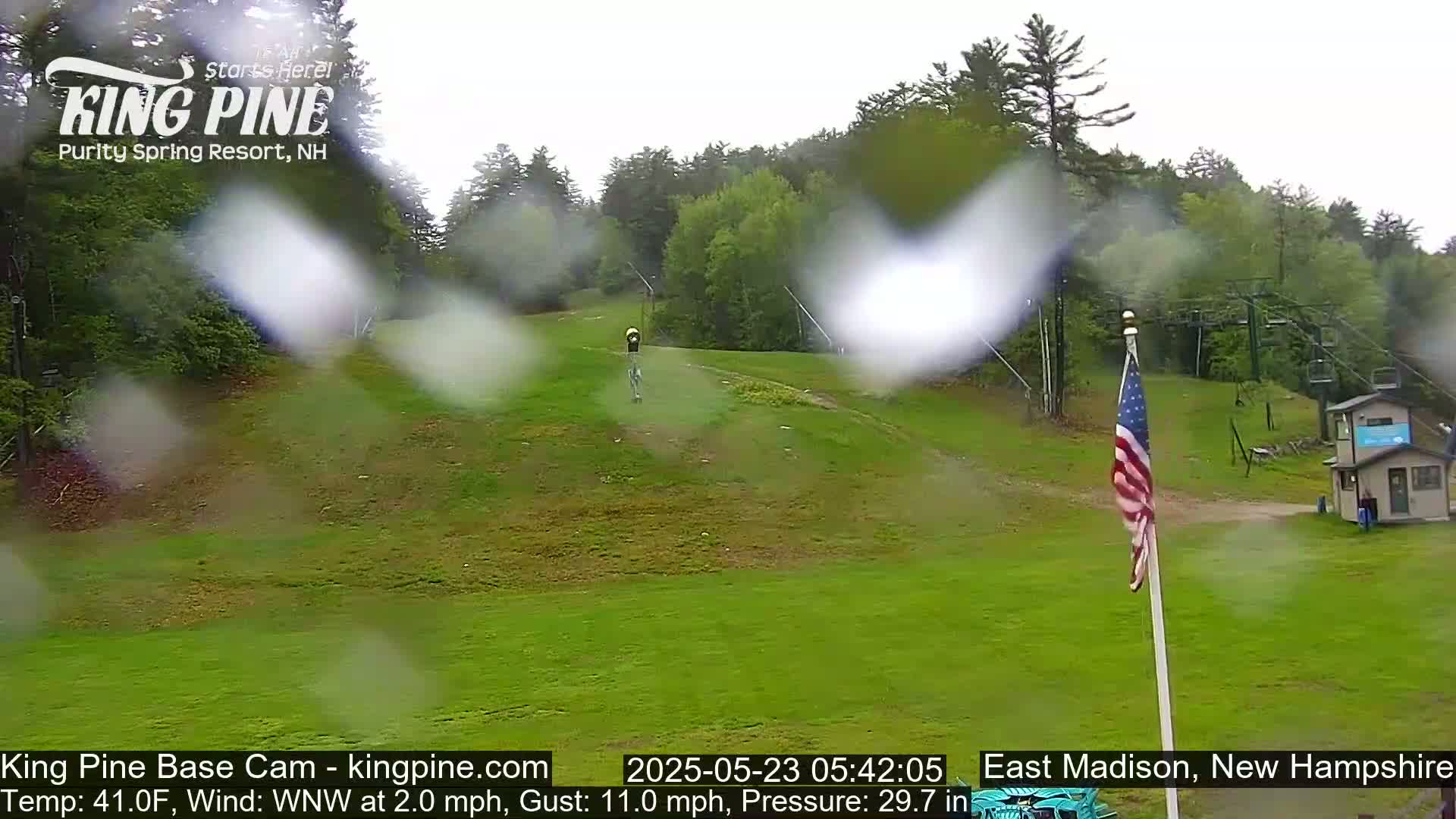 A grassy hill with a ski lift and an American flag is visible through rain, with trees in the background.