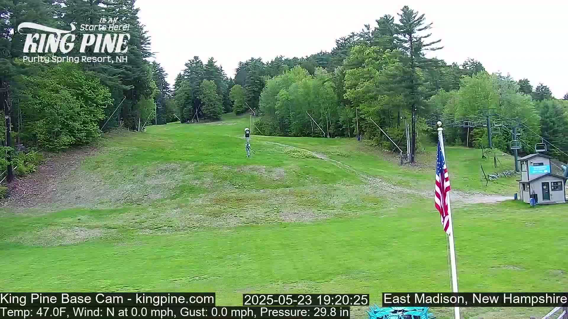 A grassy ski slope, surrounded by green trees under a cloudy sky, features a chairlift and an American flag.
