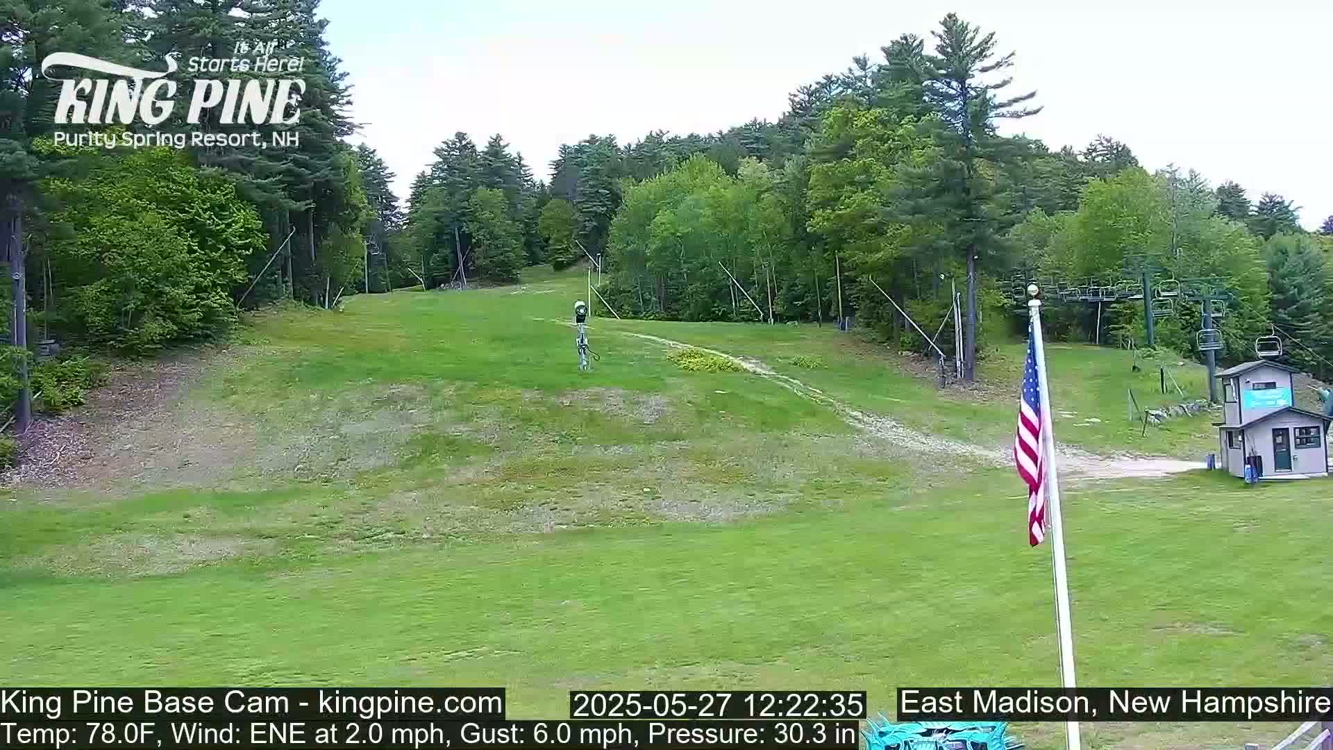 A grassy ski slope at the base of a wooded hill is shown under a partly cloudy sky, with a chairlift and American flag visible.